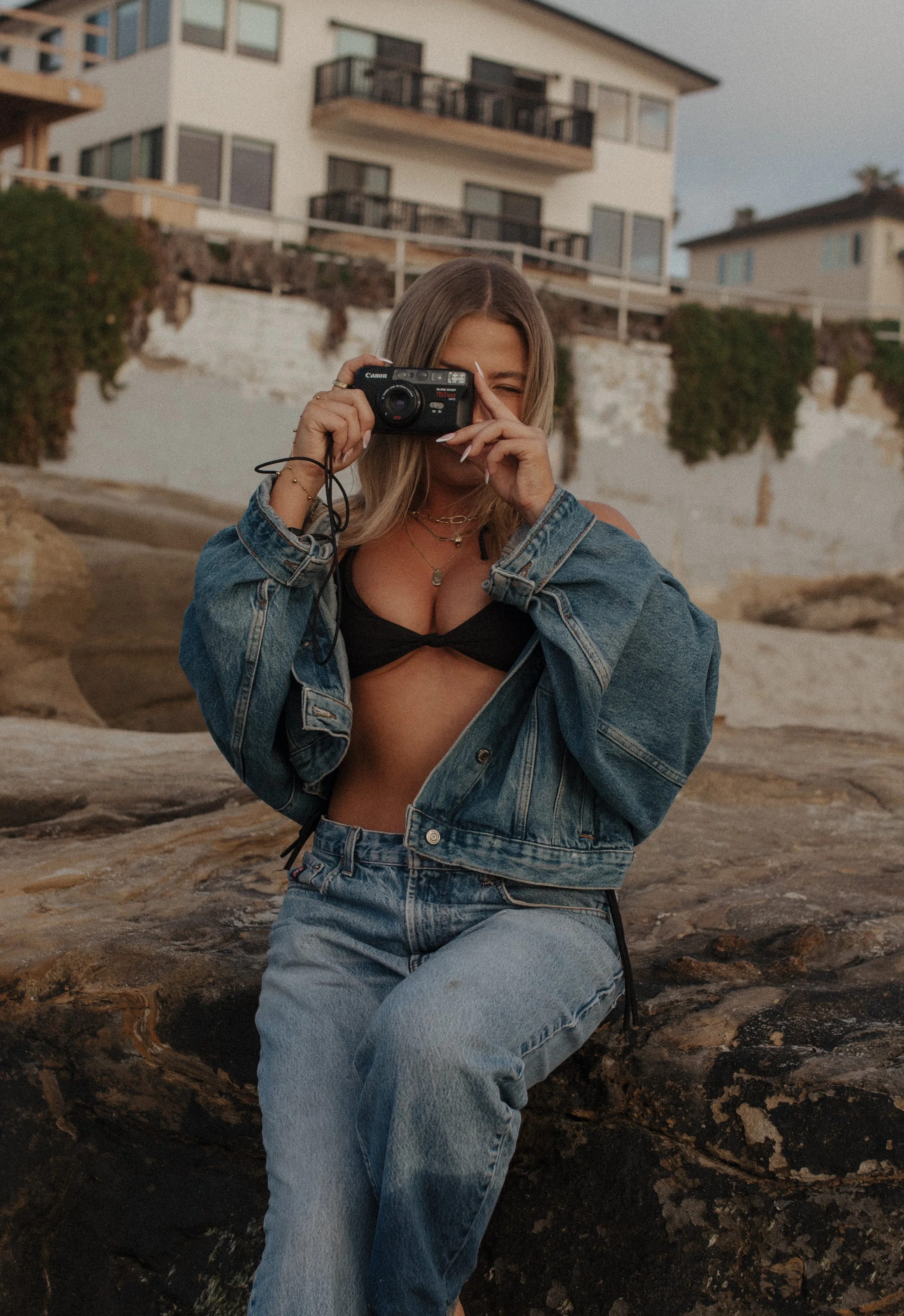 A woman wearing a denim jacket and jeans, holding a camera up to her eye, sitting on rocks near a beach, with houses in the background.