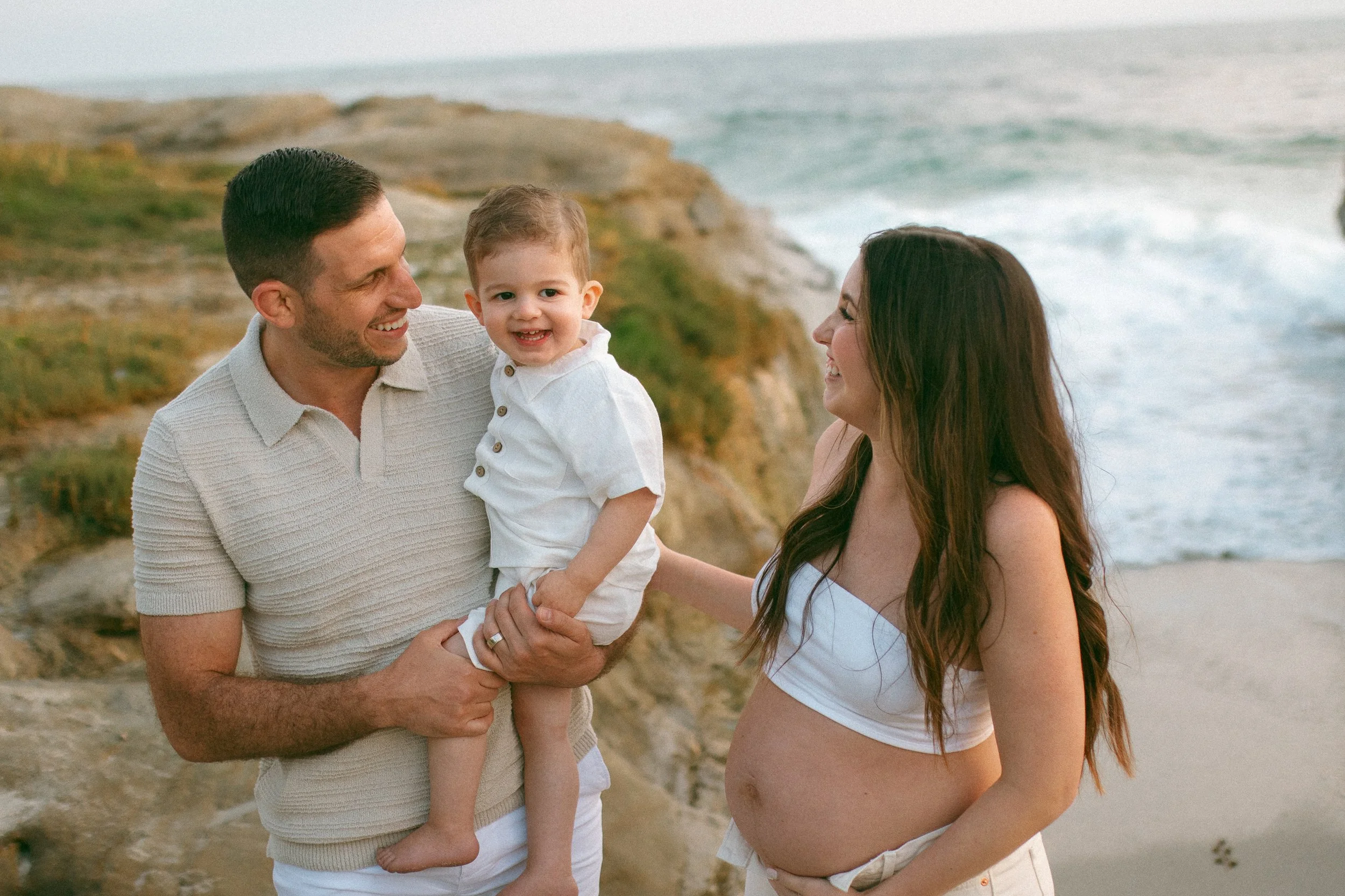 A happy family of three on the beach, with a pregnant woman, a man, and a young child, smiling at each other.
