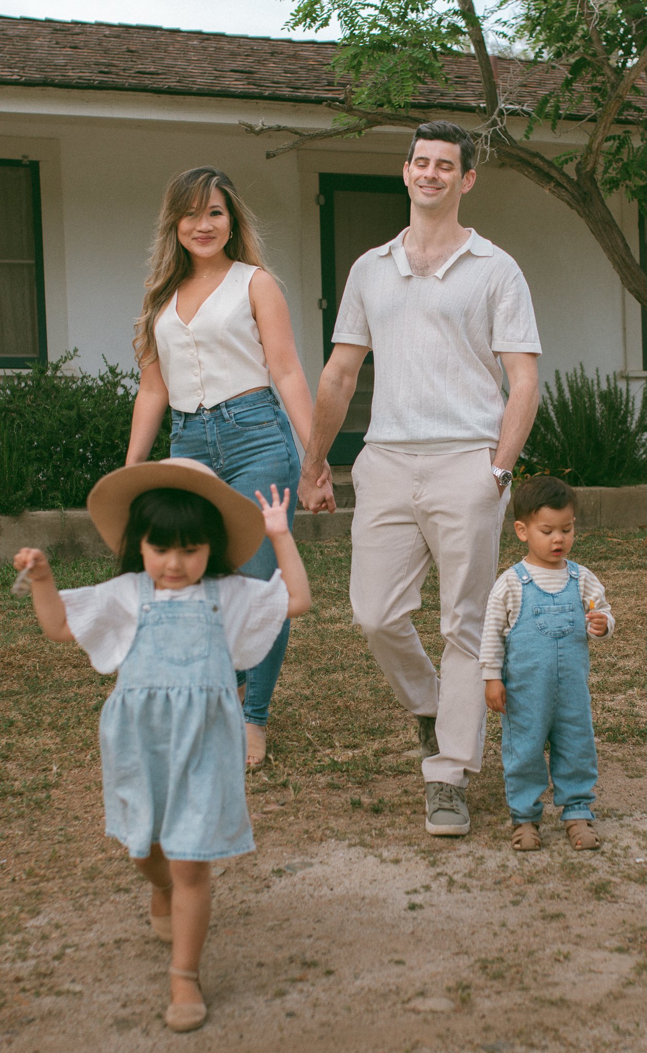 A happy family walks hand-in-hand outdoors in front of a house, with two young children. The mother and father are smiling, and the children are playful, one wearing a sun hat and the other holding a small object.