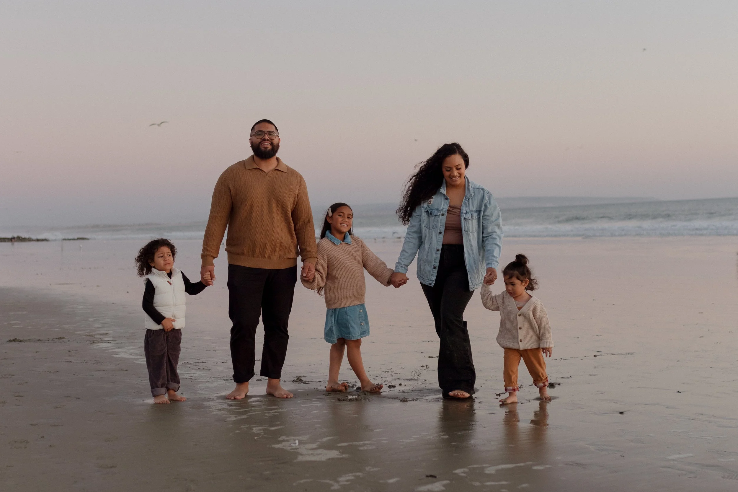 Family walking hand in hand on the beach during sunset, with the ocean in the background.