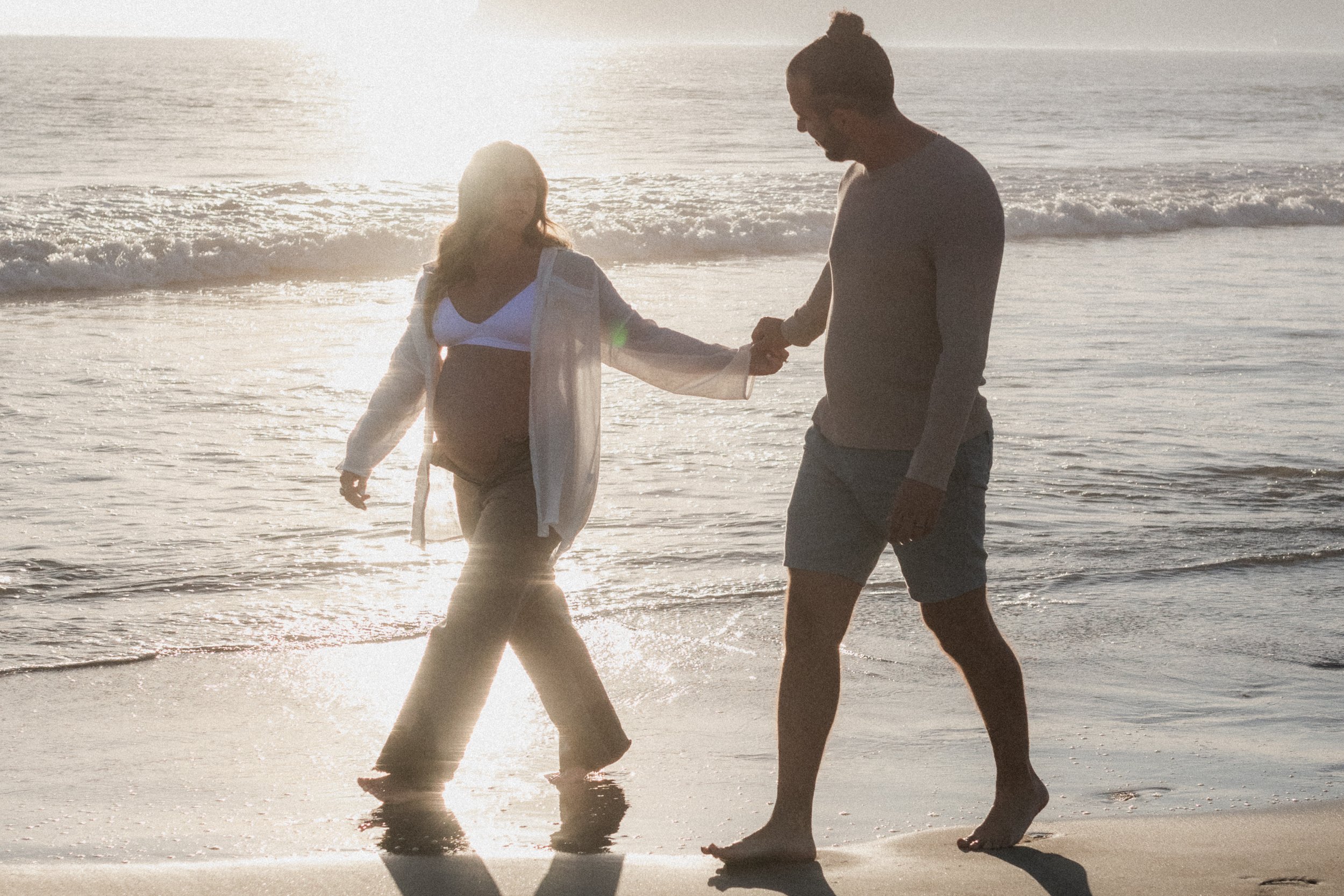 A pregnant woman and a man holding hands while walking on the beach at sunset.