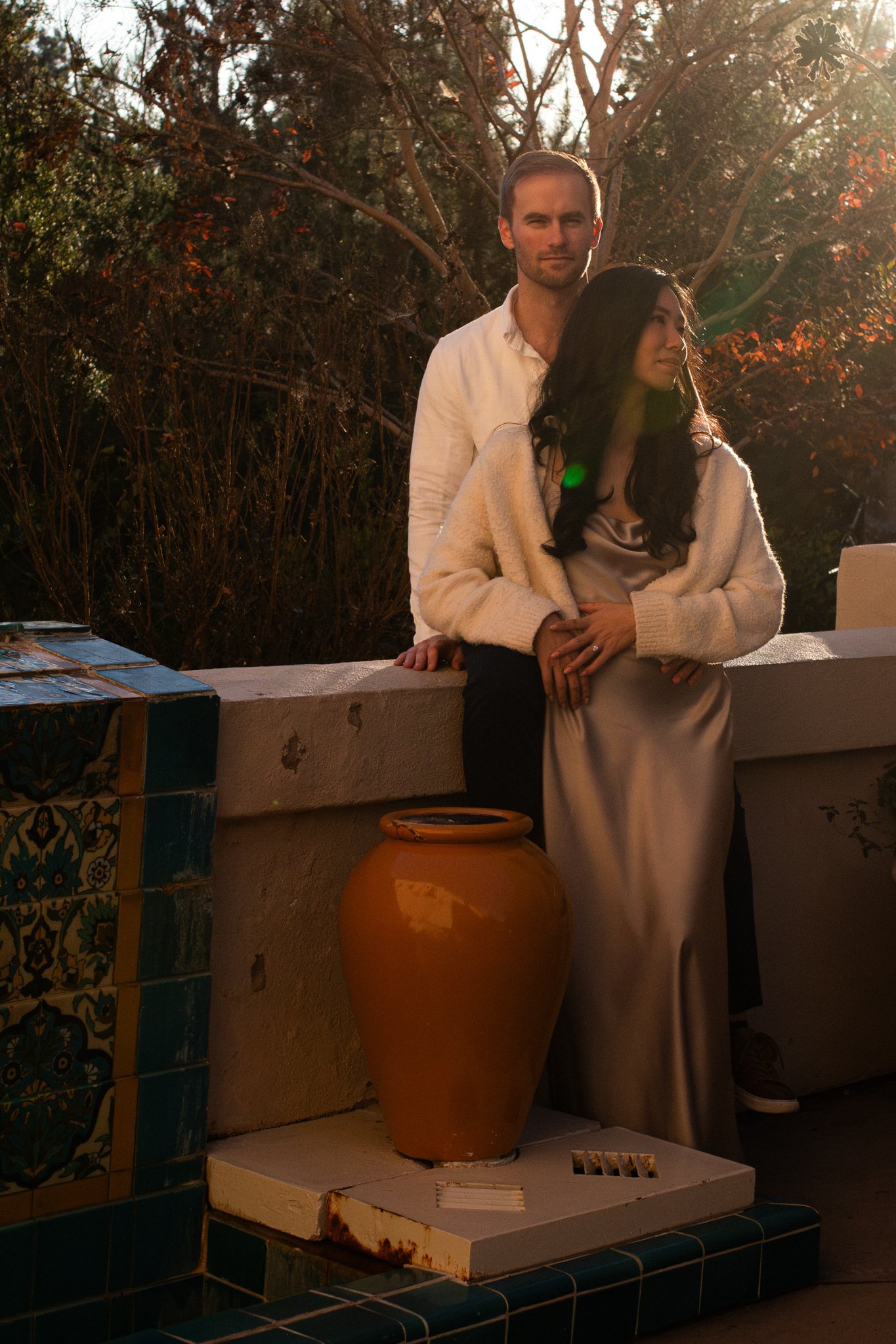 A man and woman stand outdoors on a patio during sunset, with trees in the background. The woman leans against a ledge, wearing a satin dress and cozy sweater, while the man stands behind her, dressed in a white shirt. There is a large orange clay po