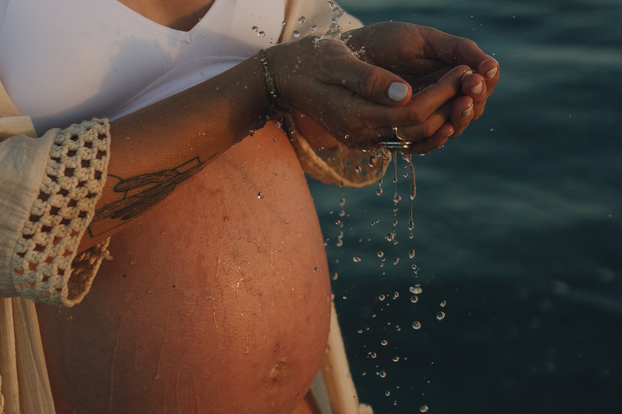 A person with a tattoo on their arm is washing their hands or holding water near their chest, with water droplets falling from their hands, near a body of water.