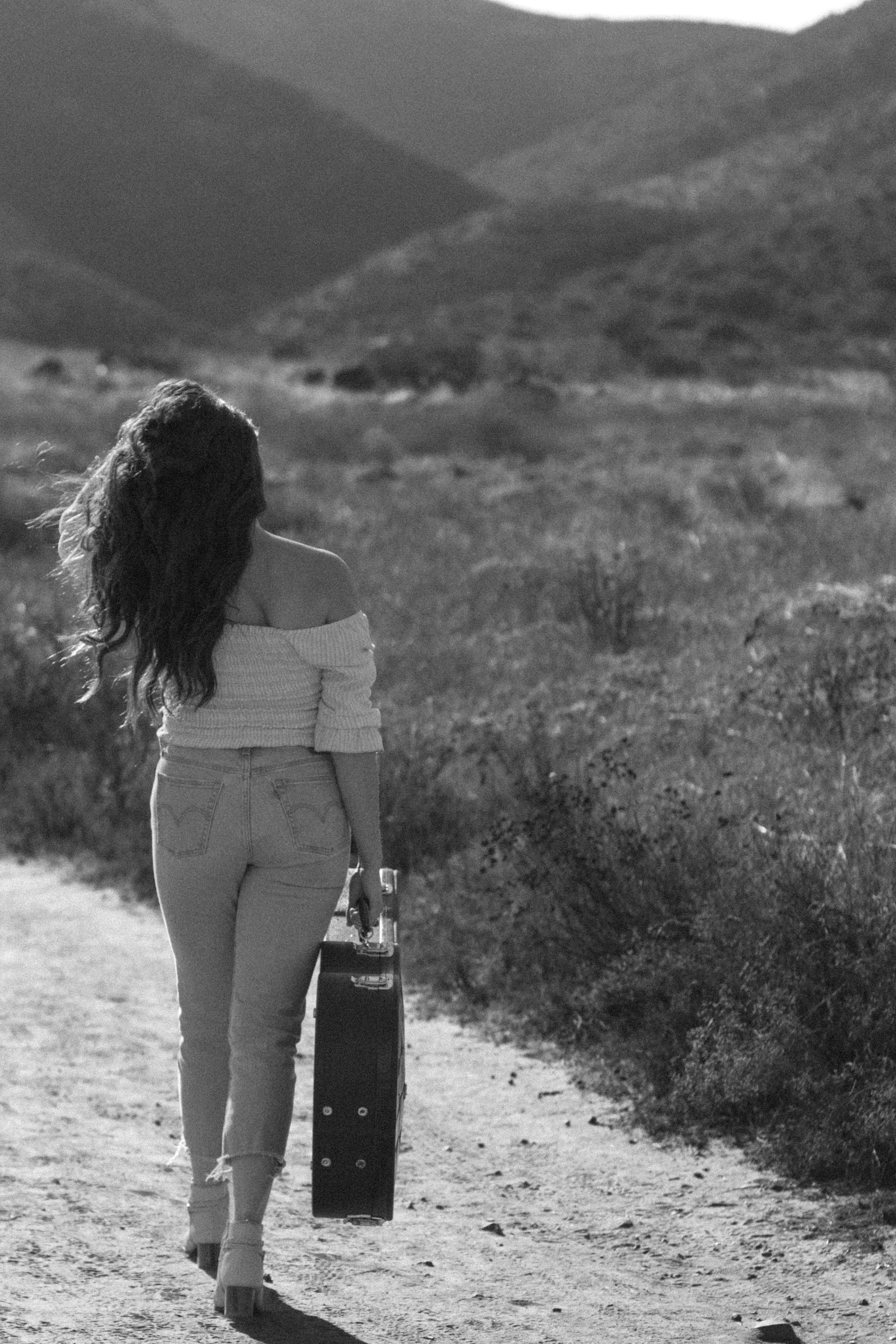 A woman walking on a dirt path in a mountainous landscape, carrying a suitcase in one hand and a glass of water in the other, dressed in an off-the-shoulder top and jeans, with long wavy hair.