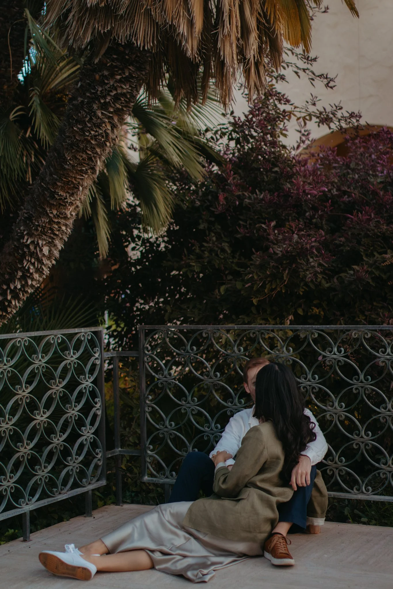 A couple sitting on the ground near a decorative metal fence, surrounded by large green plants and trees, with one person sitting behind the other.
