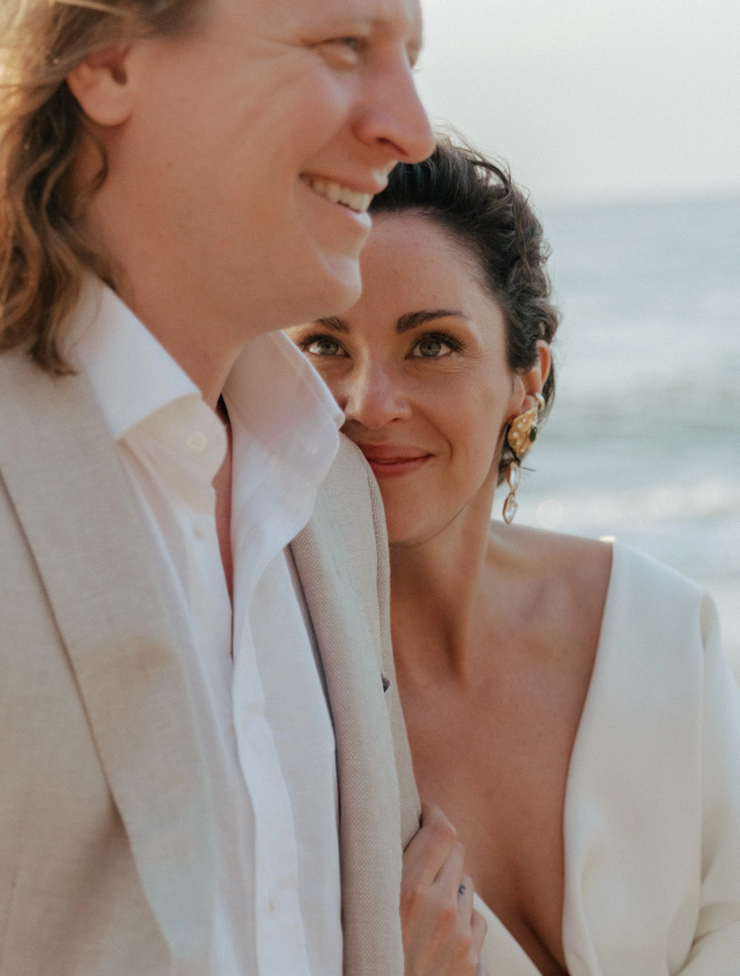 A close-up of a smiling couple at the beach, with the woman gazing at the man, who is wearing a beige blazer and a white shirt.