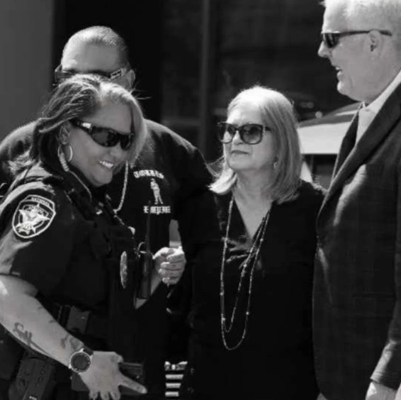 Group of four people, two women and two men, engaged in conversation outdoors. The woman on the left is in police uniform and smiling, the woman in the middle has glasses and a serious expression, and the two men are dressed in suits, with the man on the right wearing sunglasses.
