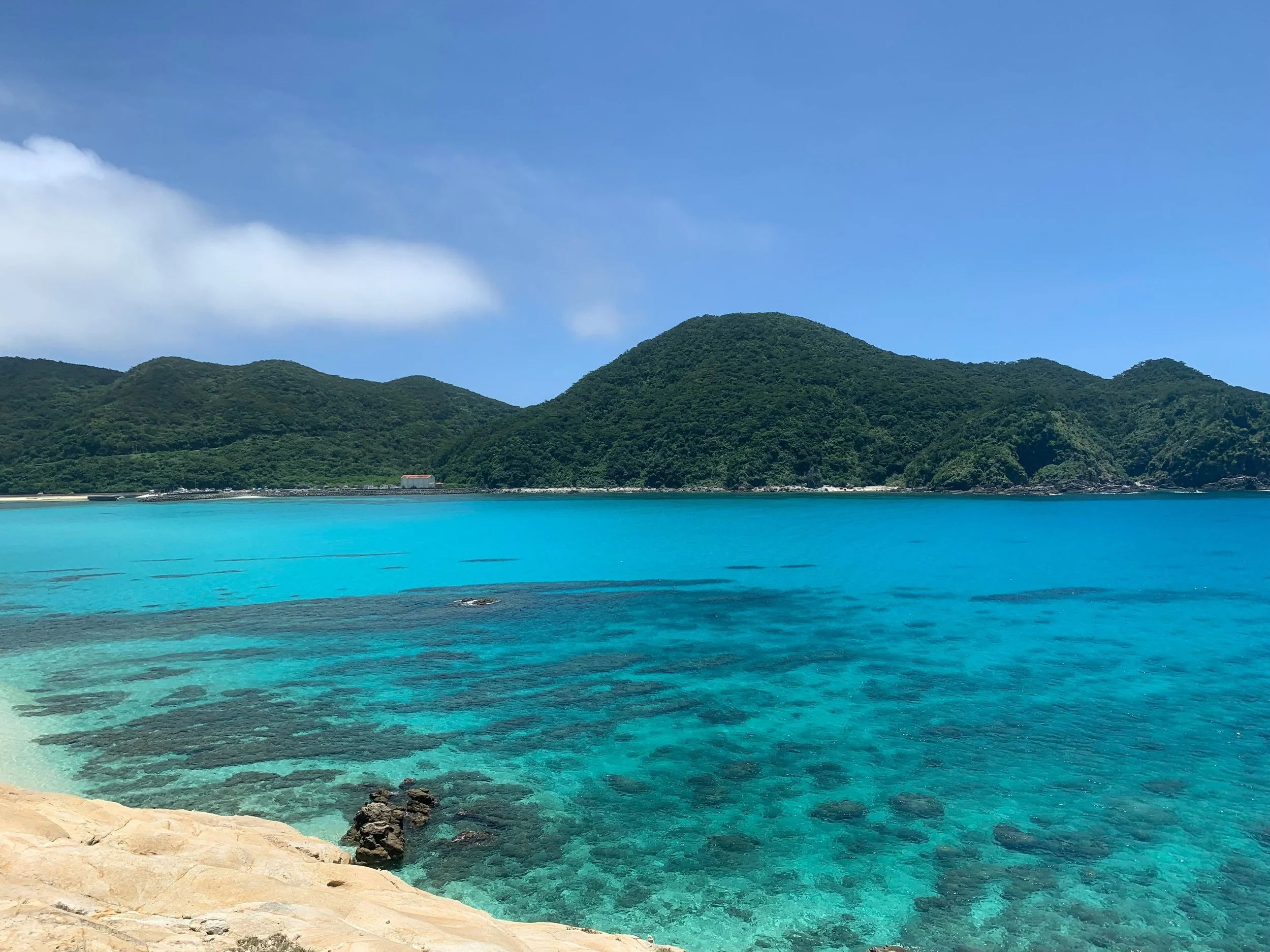Tropical ocean with clear turquoise water near a sandy shoreline, green mountainous land in the background, under a partly cloudy blue sky.