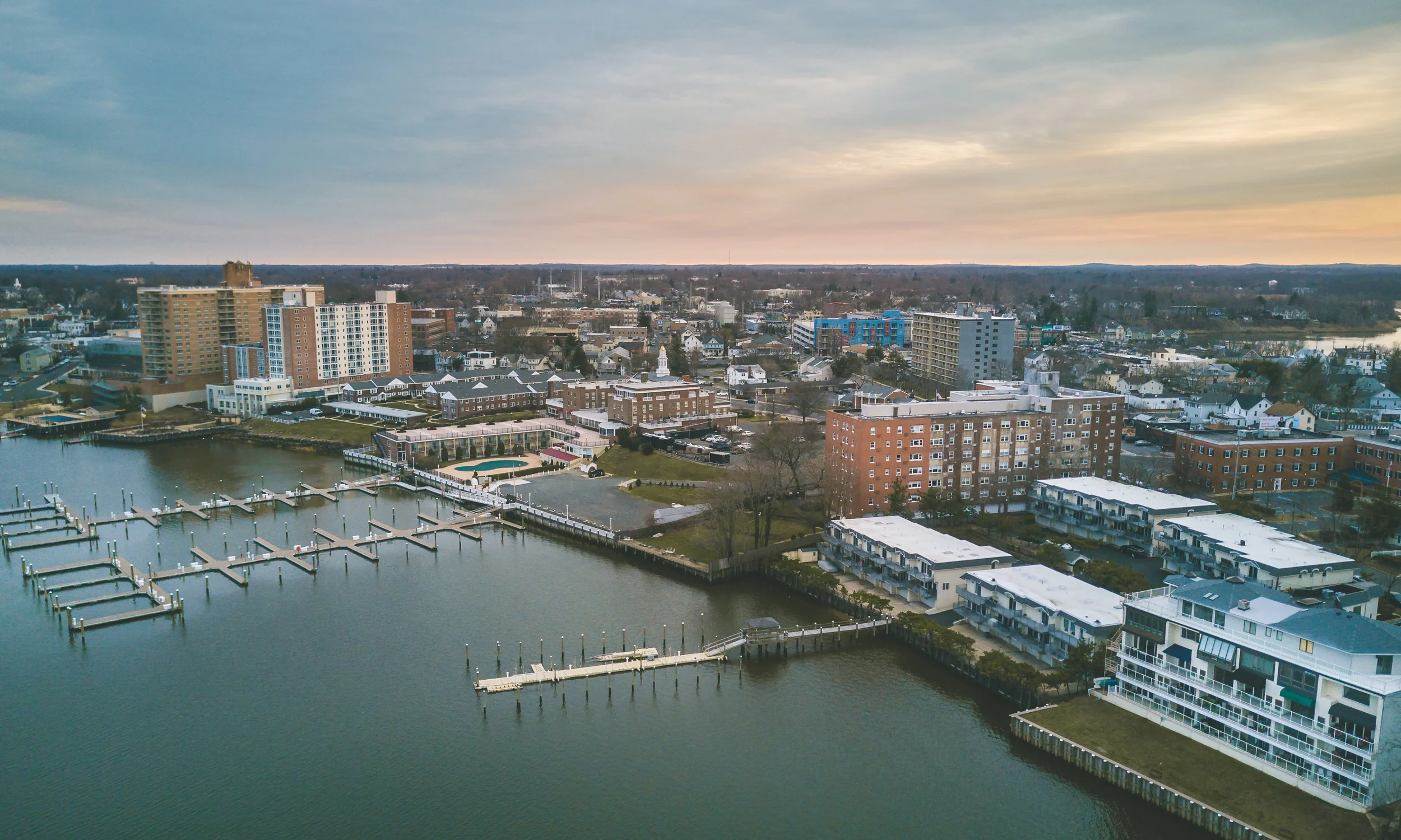 Aerial view of a city with a waterfront, featuring boats docked at piers, and buildings including high-rise apartments, hotels, and residential units, during sunset.