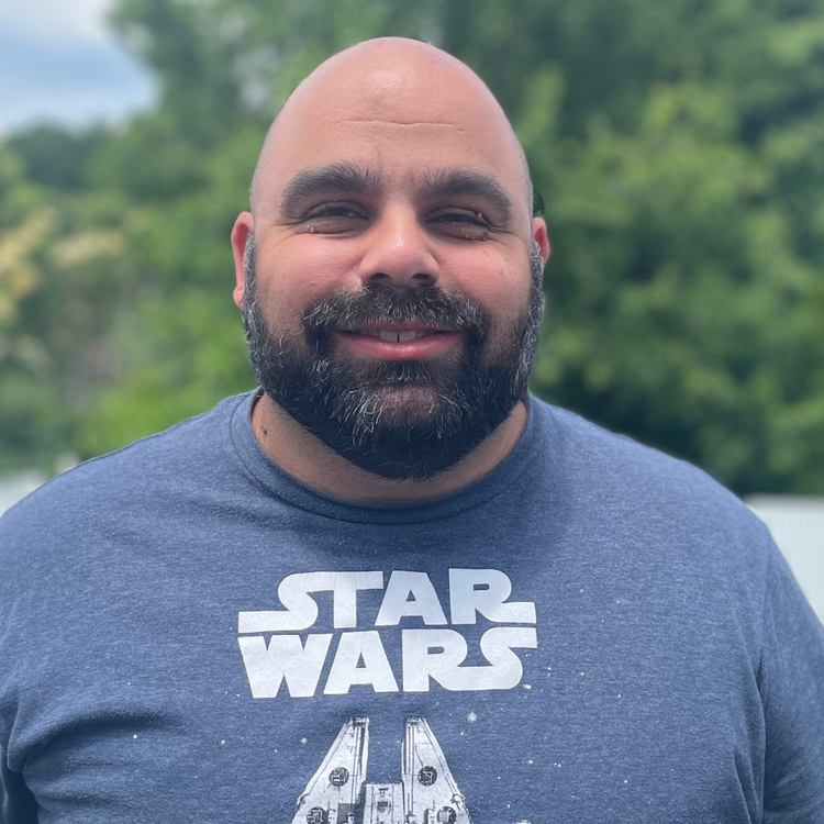 Smiling man with a beard wearing a blue Star Wars T-shirt outdoors with green trees in the background.