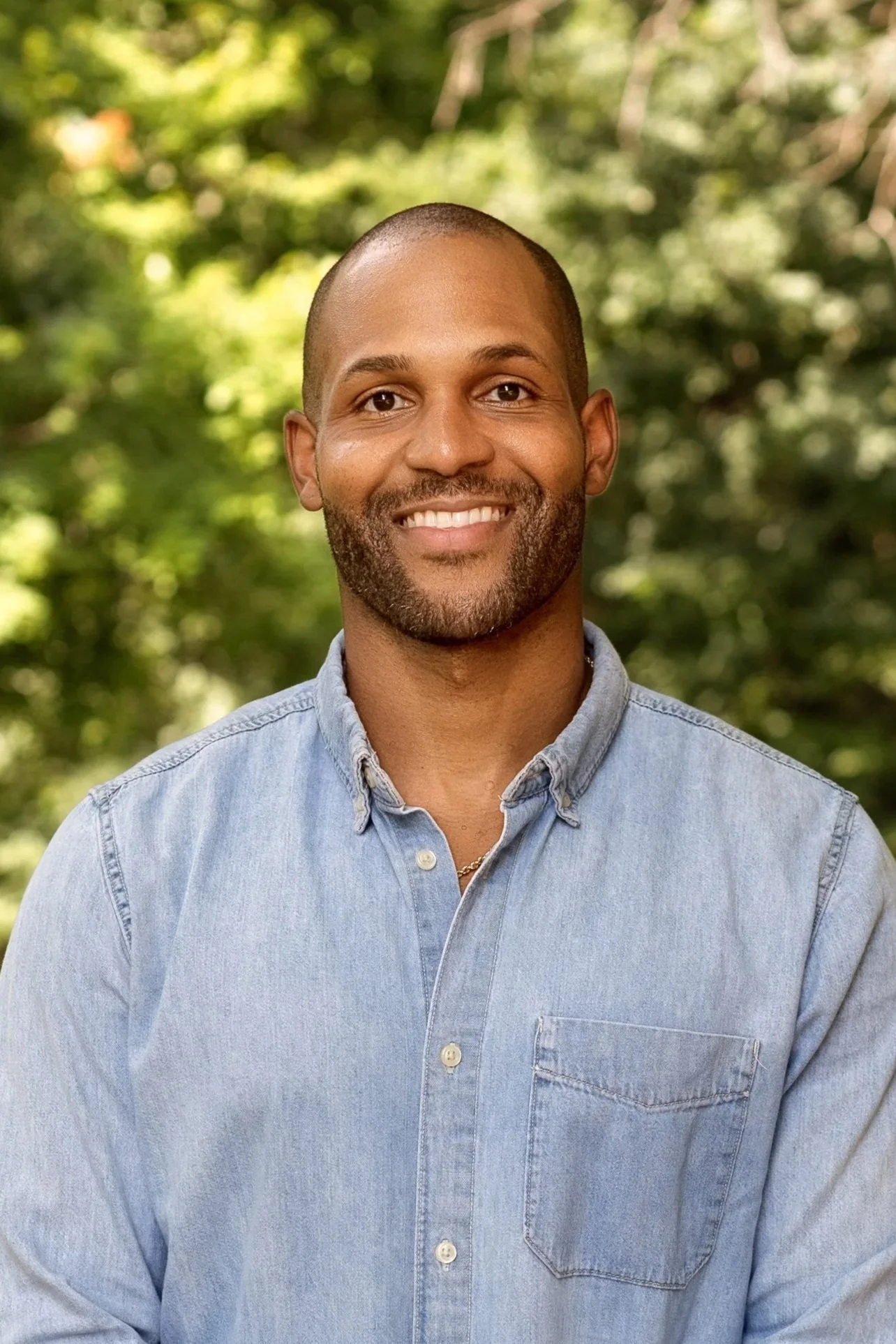 A smiling man with a beard and short hair wearing a beige sweater outdoors with trees in the background.