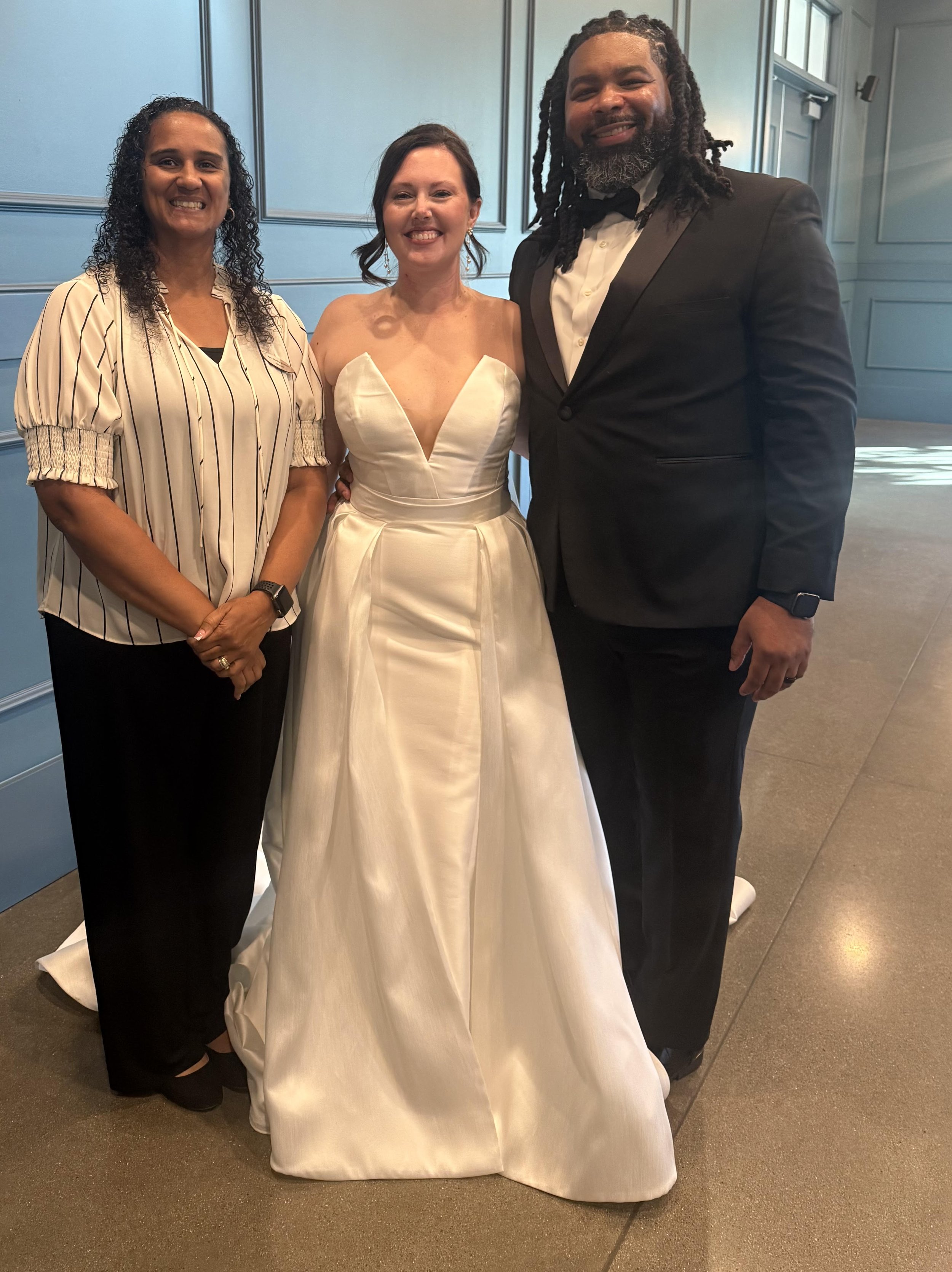 Three people, a woman in a wedding dress and a man in a tuxedo, and another woman in a striped blouse, smiling and standing close together indoors with blue walls.