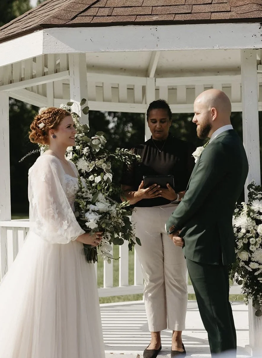 A bride and groom exchange wedding vows under a white gazebo, with an officiant standing between them and holding a tablet, surrounded by floral arrangements.