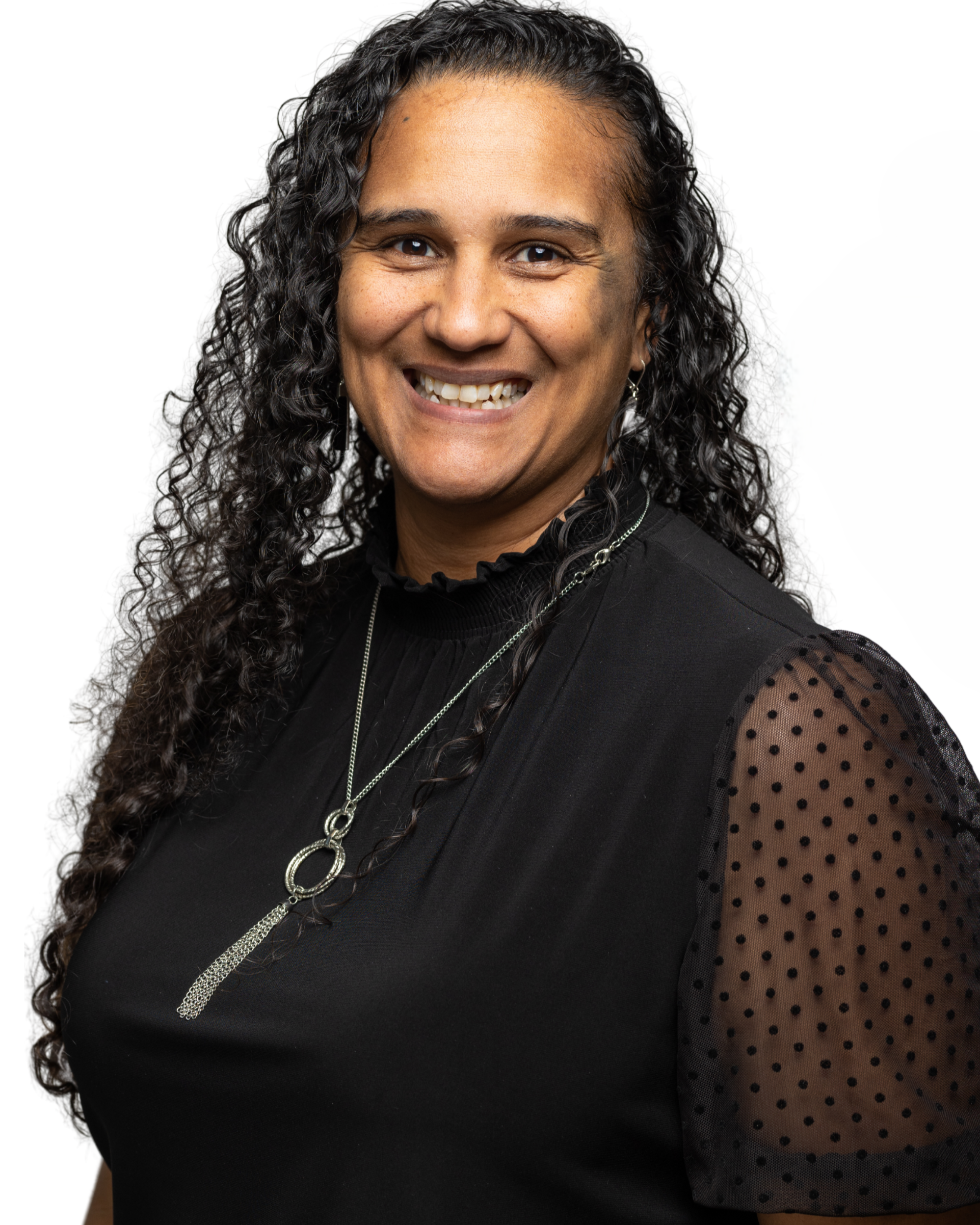 A woman with long, curly black hair smiling, wearing a black top with sheer polka-dot sleeves and a silver necklace.