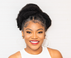 Portrait of a smiling young woman with natural hair styled in an updo, wearing a white top and hoop earrings, against a plain white background.