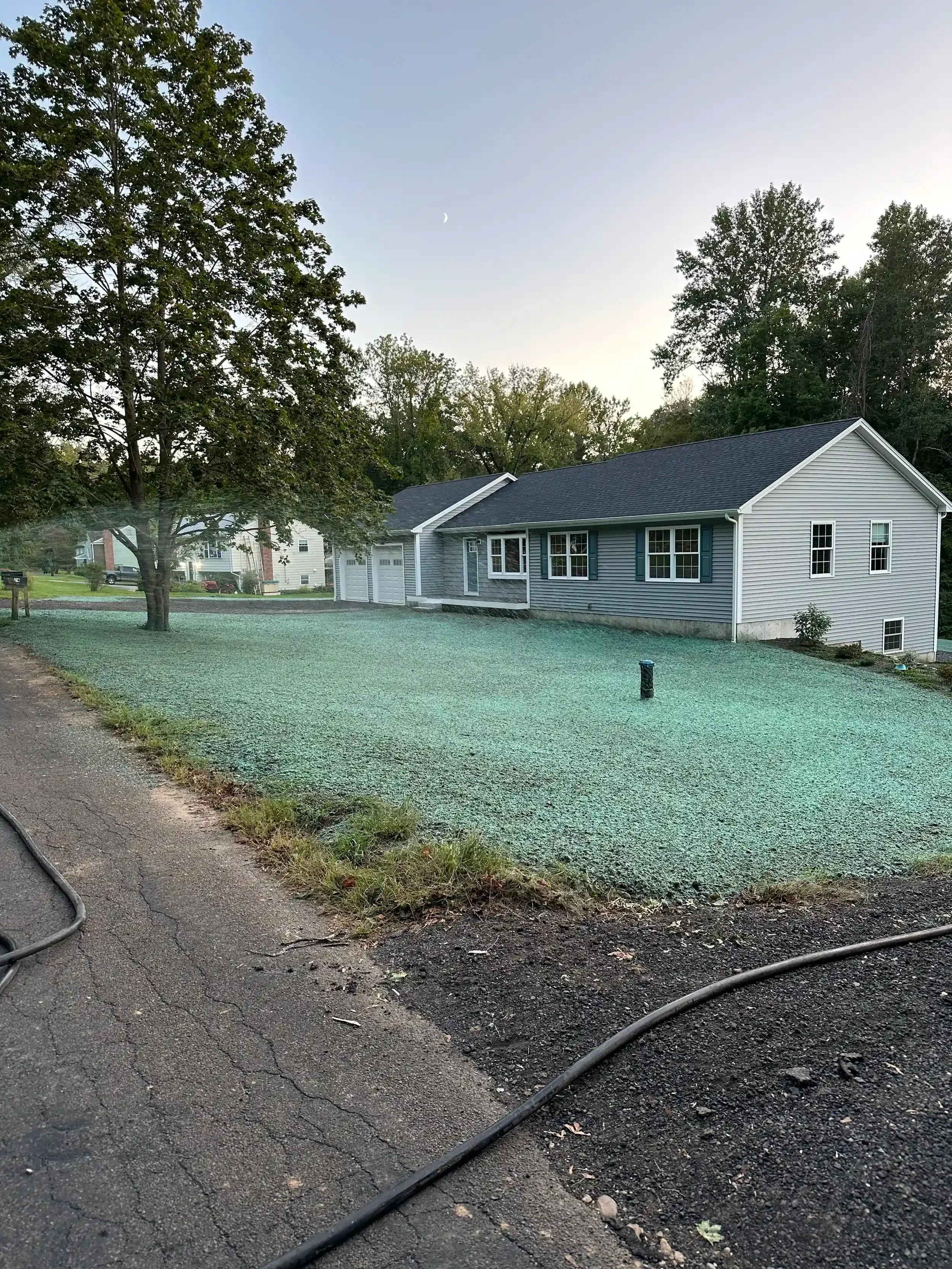 A suburban house with a gray exterior and white trim, black shutters, a two-car garage, and a grassy front yard. Tall trees are on the left side, and a small shrub is near the foundation. The sky is clear with a visible crescent moon.