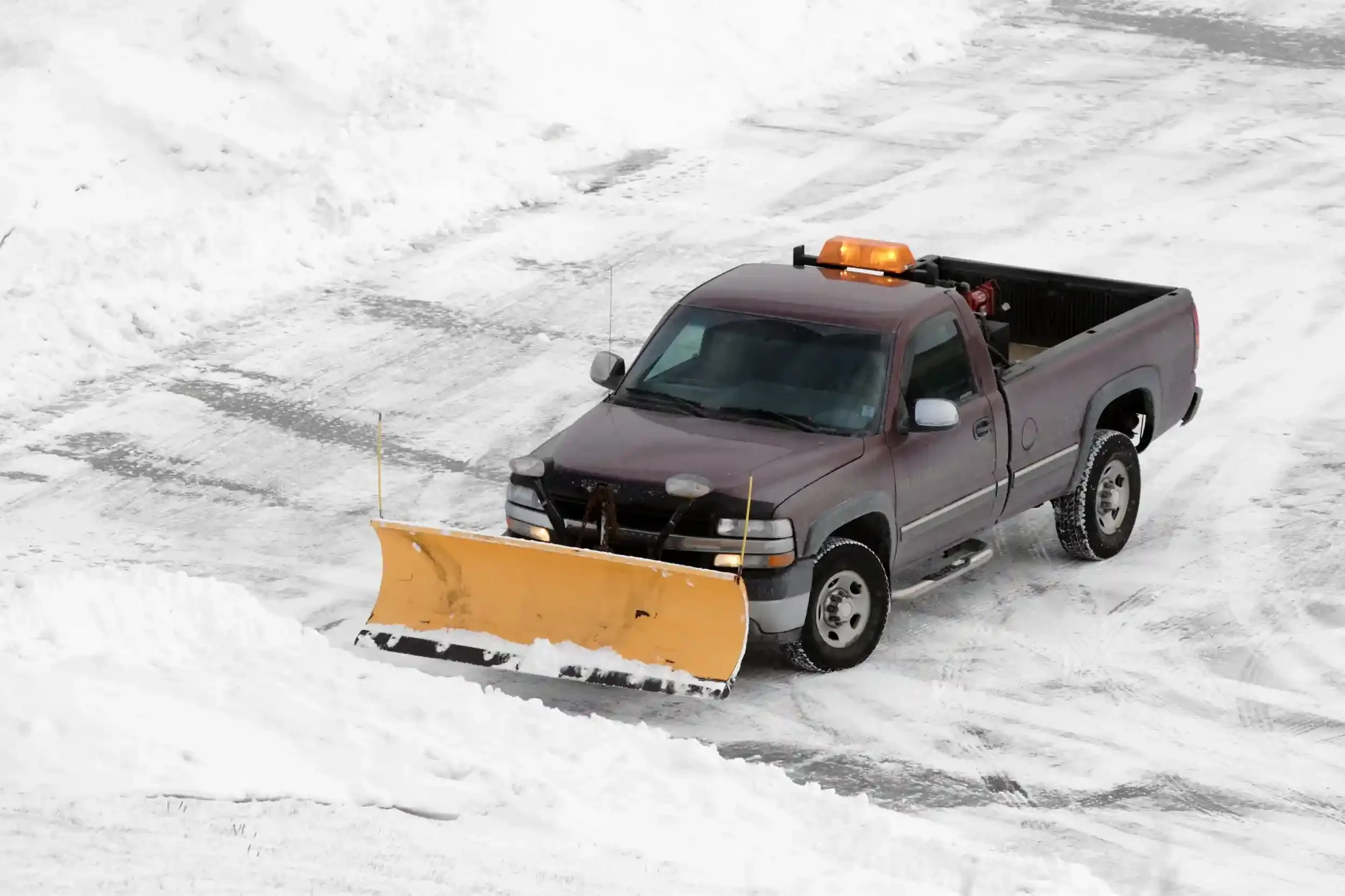 A pickup truck with a yellow snow plow attached to the front, clearing snow from a paved surface in a snowy environment.