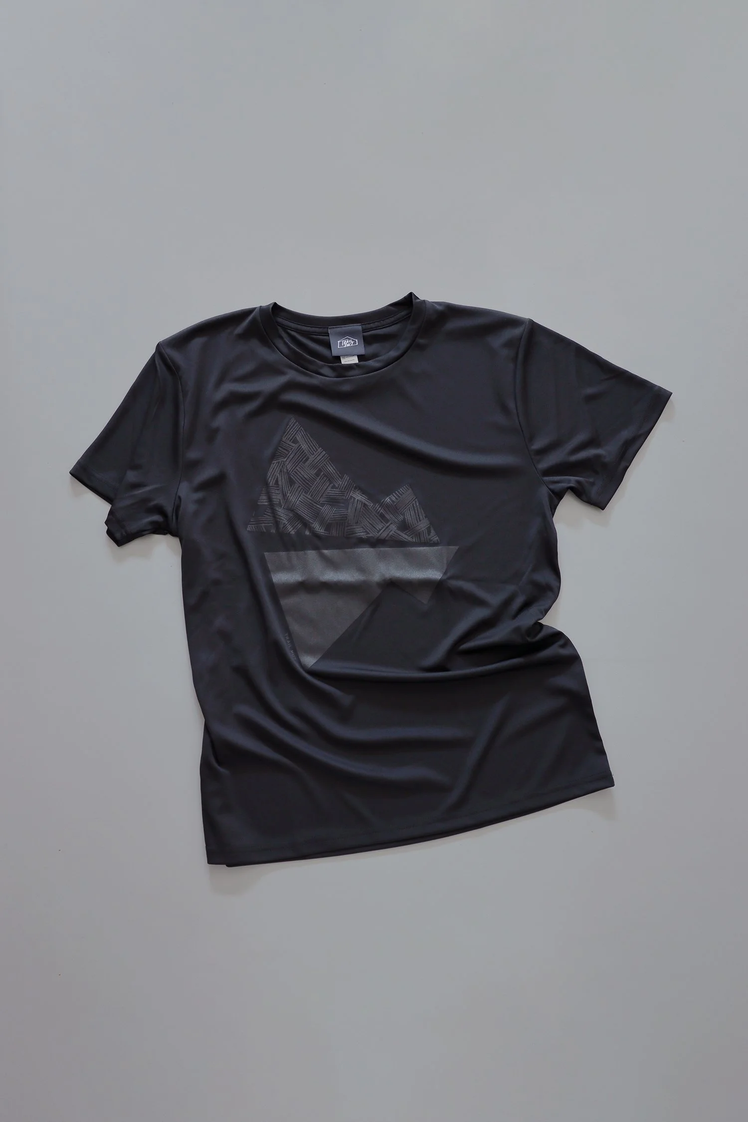 黒いベースにトーン・オン・トーンで山のモチーフをプリントしたTRAIL HUTオリジナルTシャツ。光の加減で模様が浮かび上がる、控えめでグラフィカルなデザイン。A black TRAIL HUT original T-shirt featuring a tonal mountain motif print. The subtle graphic reveals itself with light reflection, creating a refined and understated des