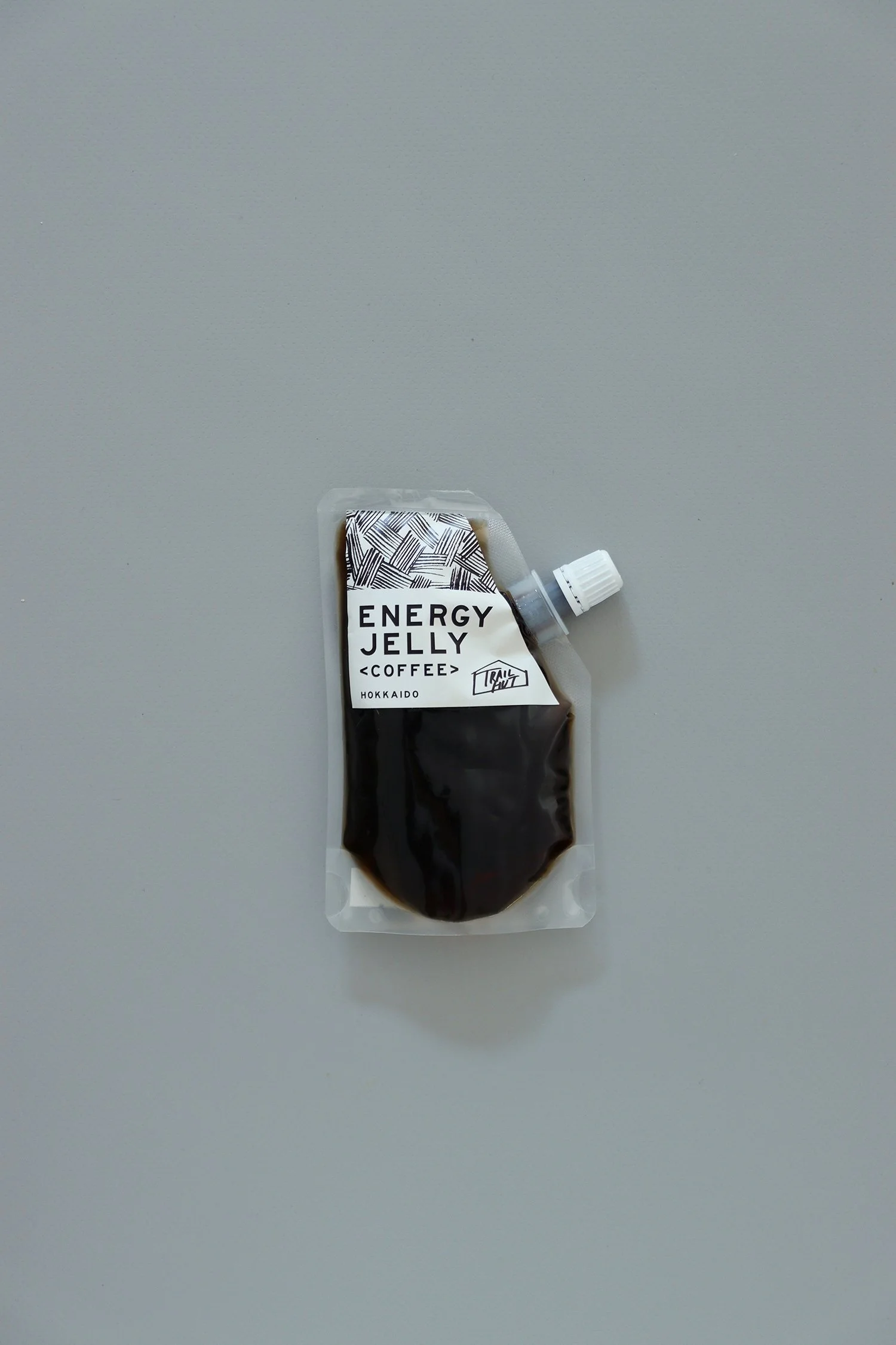 グレーの背景に置かれた、TRAIL HUTのオリジナル商品「ENERGY JELLY〈COFFEE〉」。透明パウチに黒いゼリーが入り、幾何学模様のキャップ部分とシンプルなラベルが印象的。A single pack of TRAIL HUT’s original “ENERGY JELLY ” placed on a gray background. The clear pouch reveals the dark jelly inside, with a geometric patterned