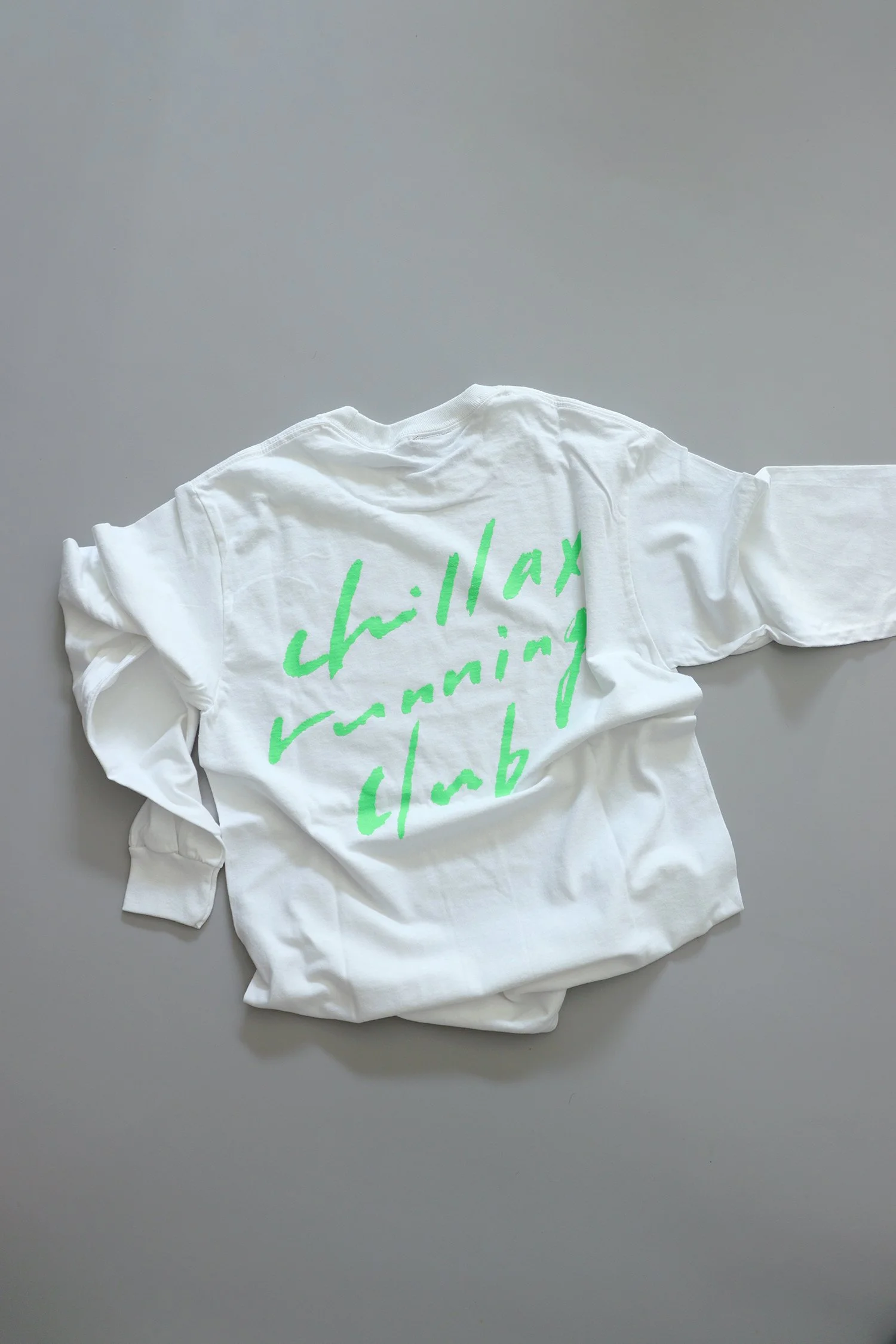 背面に「chillax running club」の手描き風文字を鮮やかなグリーンで大きく配置した、TRAIL HUTオリジナルのホワイトロングスリーブTシャツ。リラックス感とスポーティさを掛け合わせたデザイン。A white long-sleeve TRAIL HUT T-shirt with a bold handwritten-style text “chillax running club” printed in bright green across the back. A desig