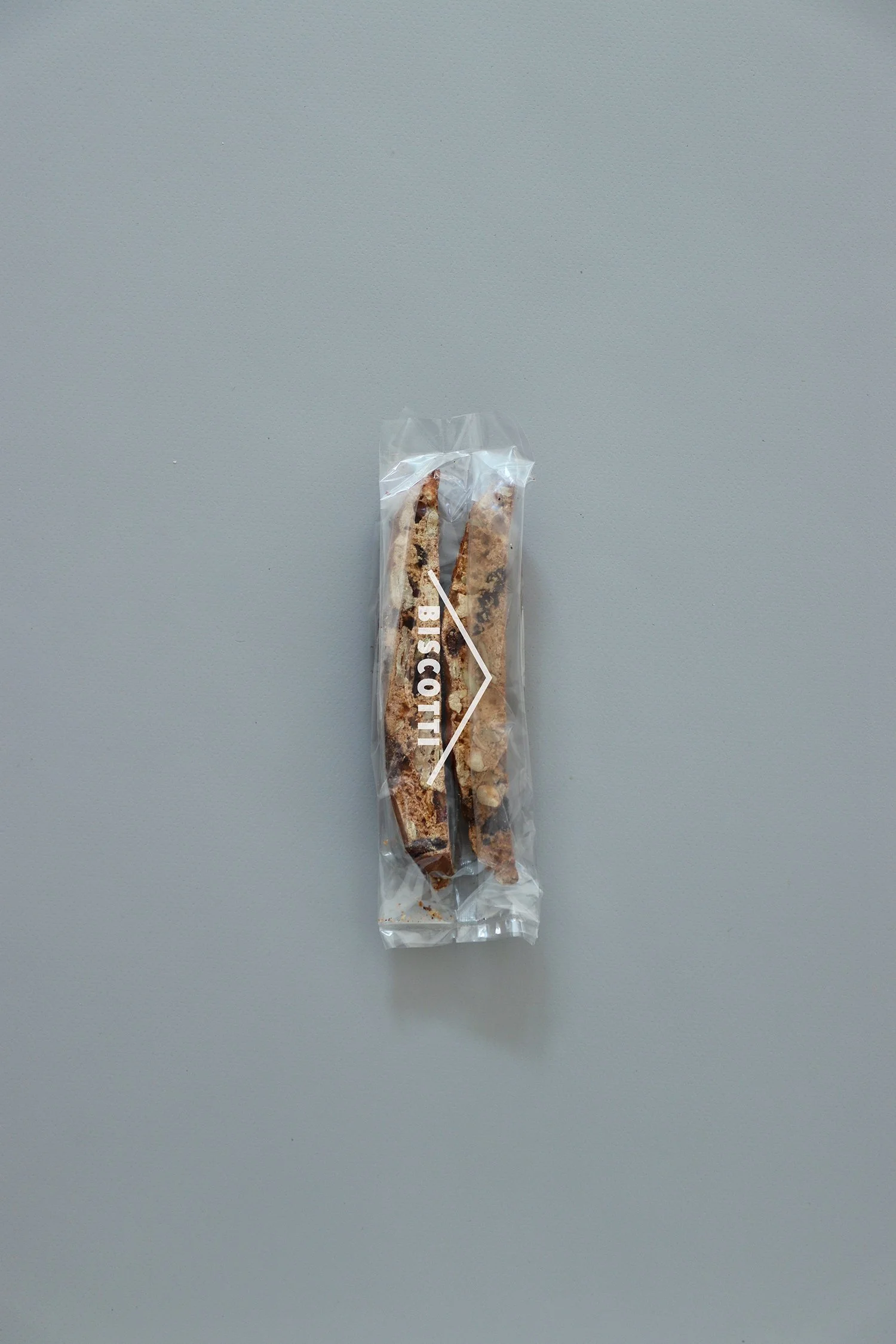 グレーの背景に置かれた、TRAIL HUTのオリジナル「BISCOTTI」。透明の細長いパッケージに2本のビスコッティが入り、表面にはシンプルなロゴがプリントされている。TRAIL HUT’s original “BISCOTTI” on a gray background. The slim clear package contains two biscotti sticks, with a simple white logo printed across the front.