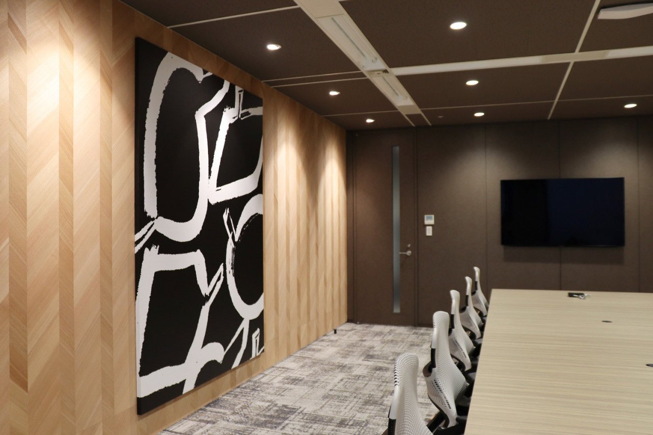 NTT-AT会議室の壁に設置された大判の抽象アート。黒地に白の手描き風モチーフが大胆に描かれている。Large abstract artwork in black and white on the meeting room wall of NTT-AT. The hand-drawn style composition adds bold contrast to the wood paneling.
