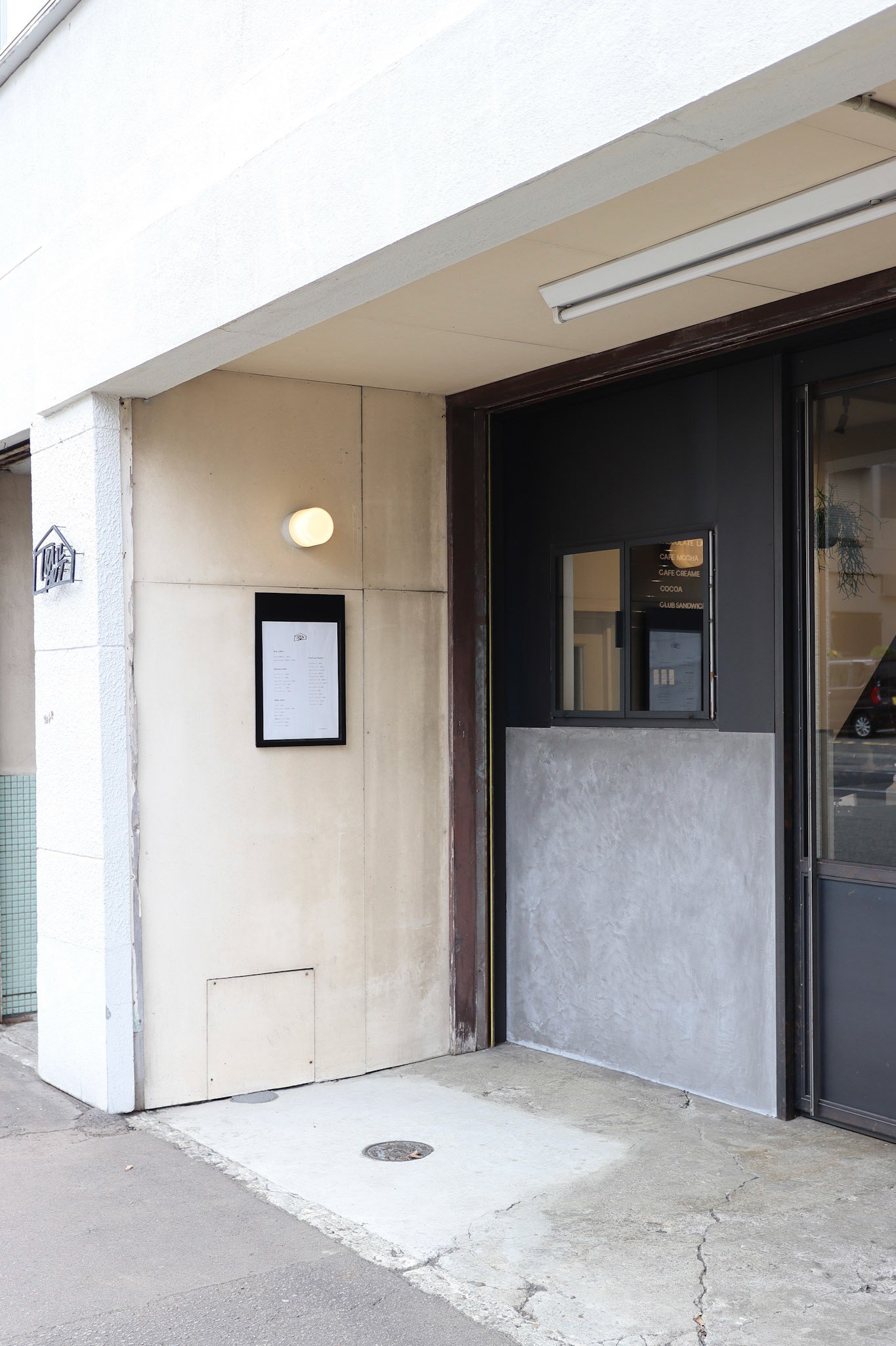 コンクリート壁とダークグレーの扉が印象的なカフェの外観。照明とメニュー表示がシンプルに配置されている。
Exterior of a café featuring a concrete wall and dark grey door, with understated lighting and neatly framed menu display.