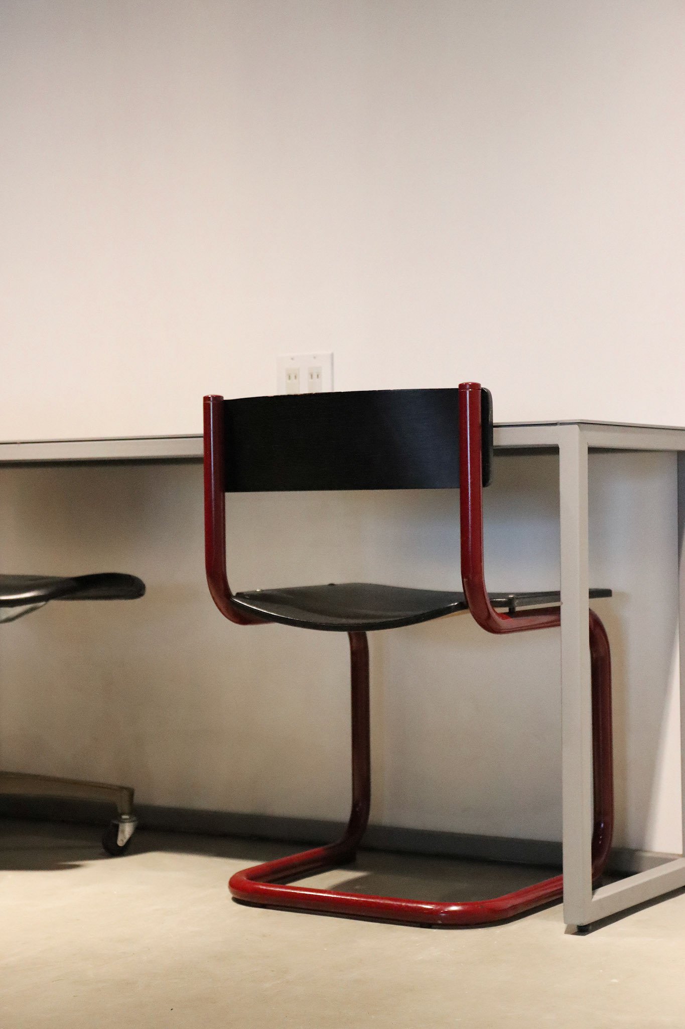 グレーの作業机に合わせた黒い座面と背の赤いスチールチェア。無機質な空間に映えるアクセント。
A red steel-frame chair with black seat and backrest placed at a gray work desk, adding a bold accent to the minimalist space.