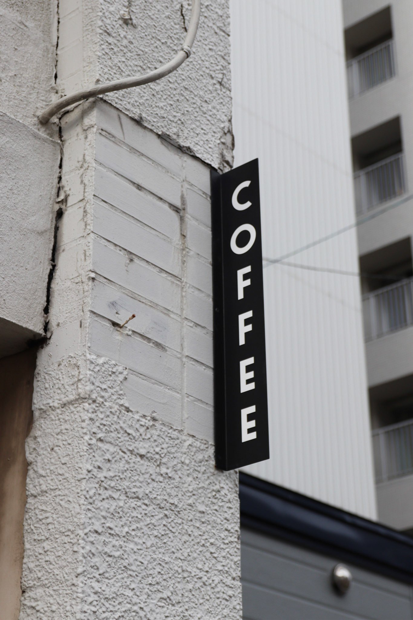 ザラついた質感の白い壁に取り付けられた黒地に白文字の縦型「COFFEE」サイン。
A vertical “COFFEE” sign with white letters on a black background, mounted on a textured white exterior wall.