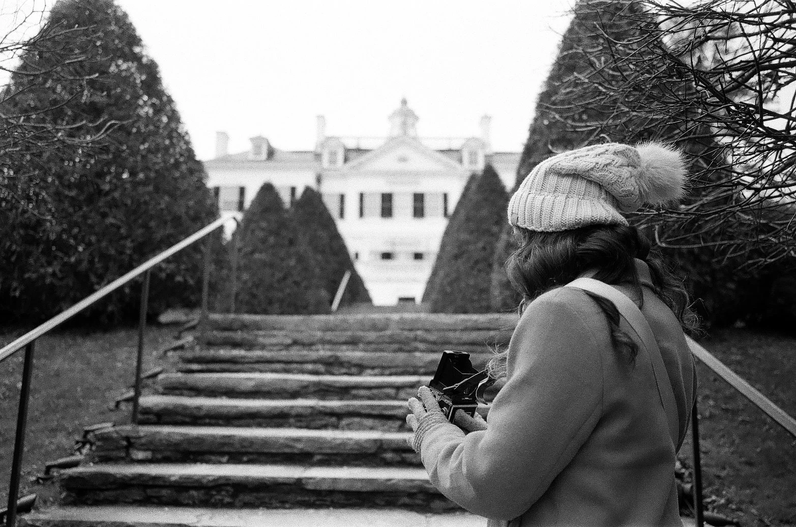 A woman wearing a knit hat and coat is taking a photo with a camera on a set of outdoor stairs. There are trees and a large mansion in the background.