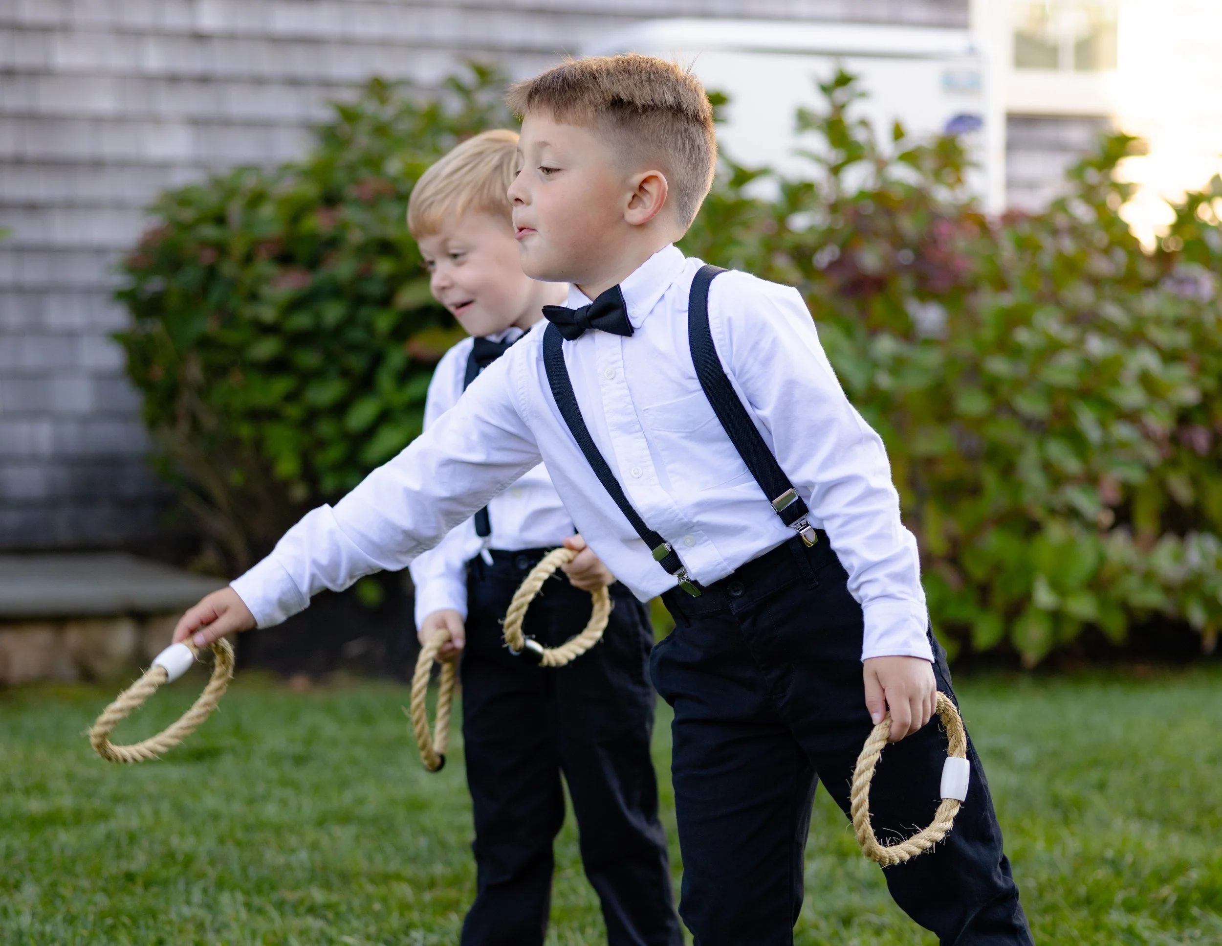 Two young boys dressed in white shirts, black pants, and suspenders, playing ring toss on a grass lawn outdoors during daytime.