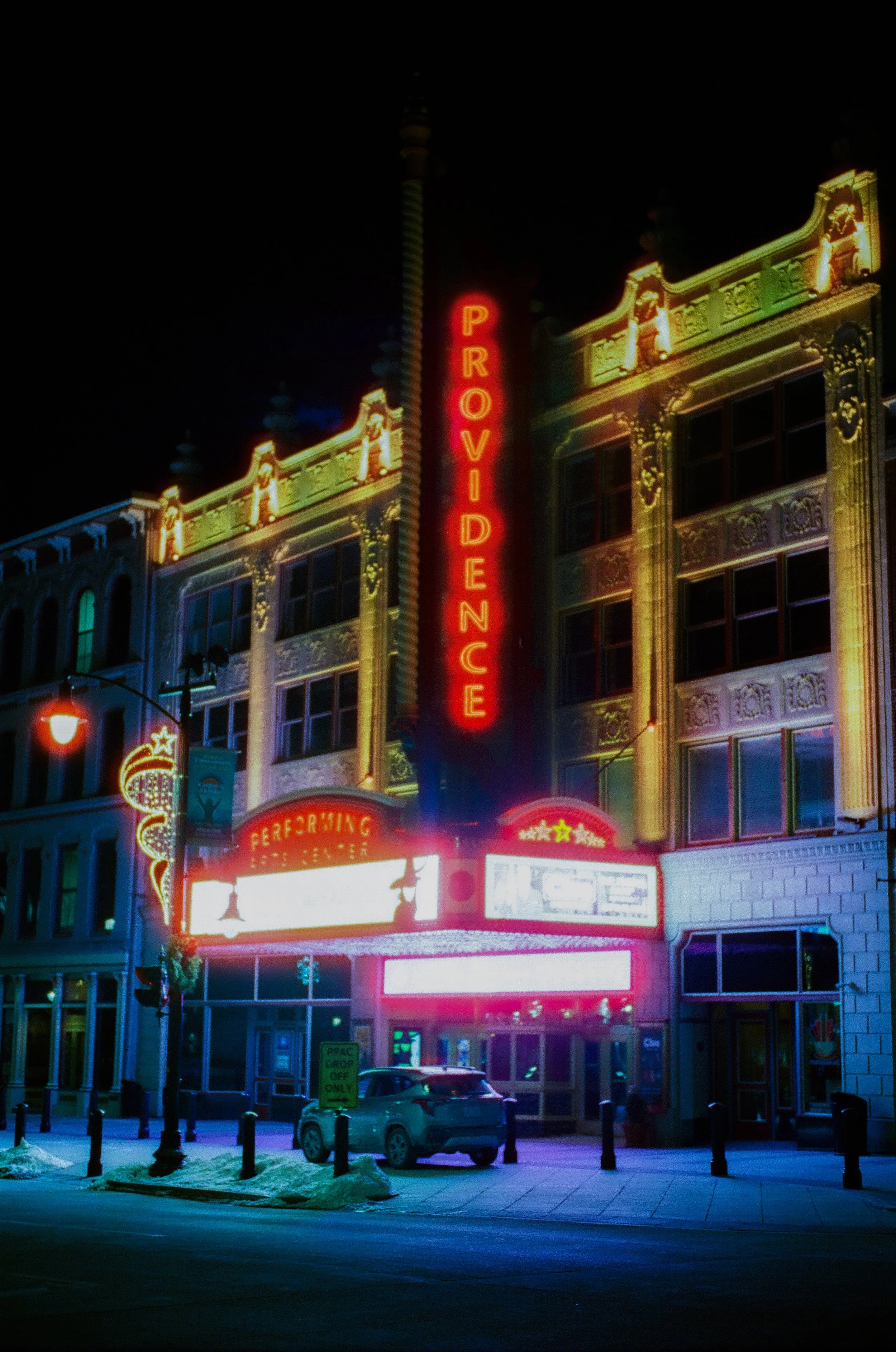 Neon-lit theater marquee and signage at night, with a car parked in front, snow on the ground, and ornate building facade in the background.