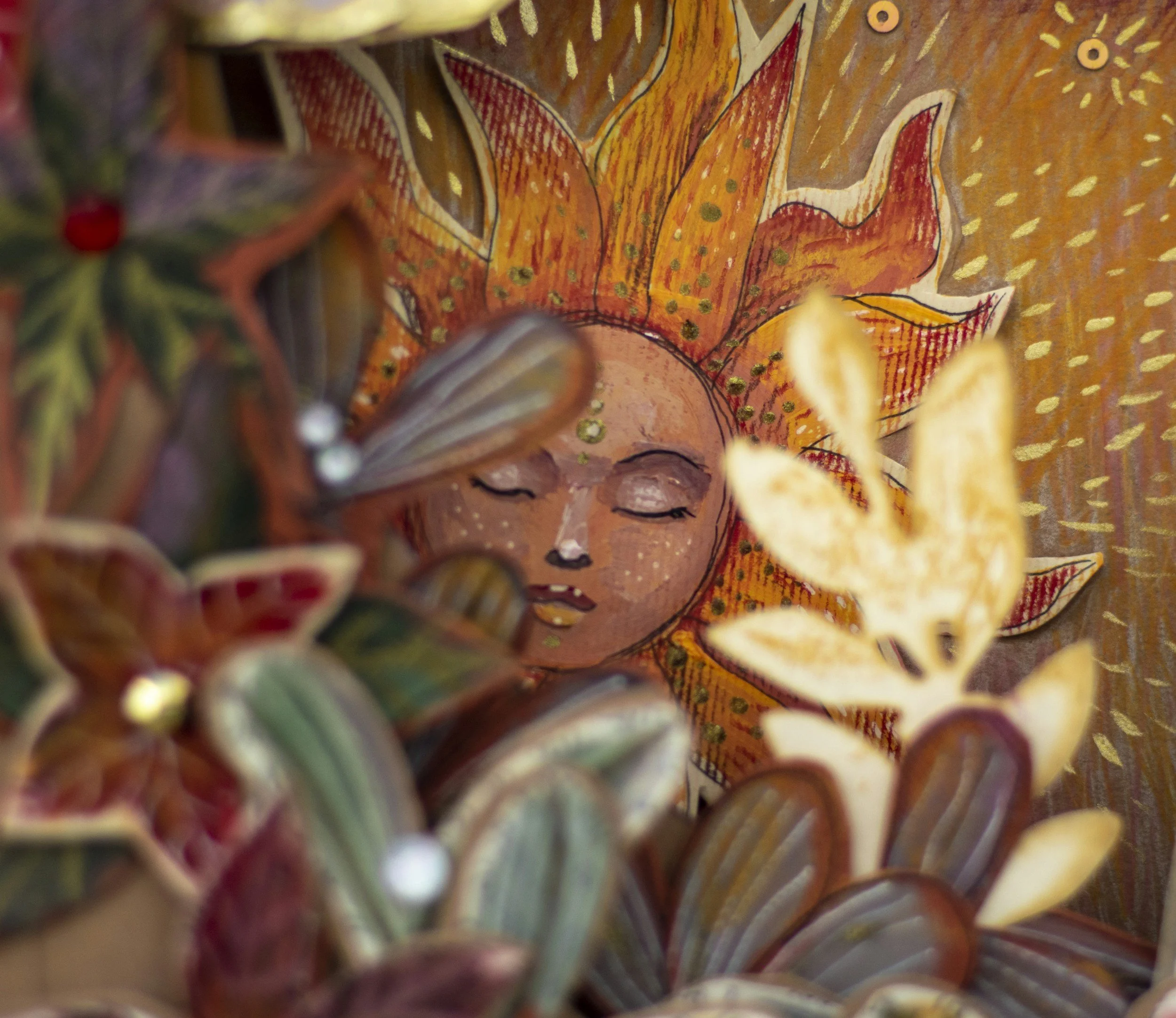 A colorful boho sun inspired wall hanging depicting a face with closed eyes and a large sunburst behind it, partially covered with decorative leaves and flowers in the foreground.