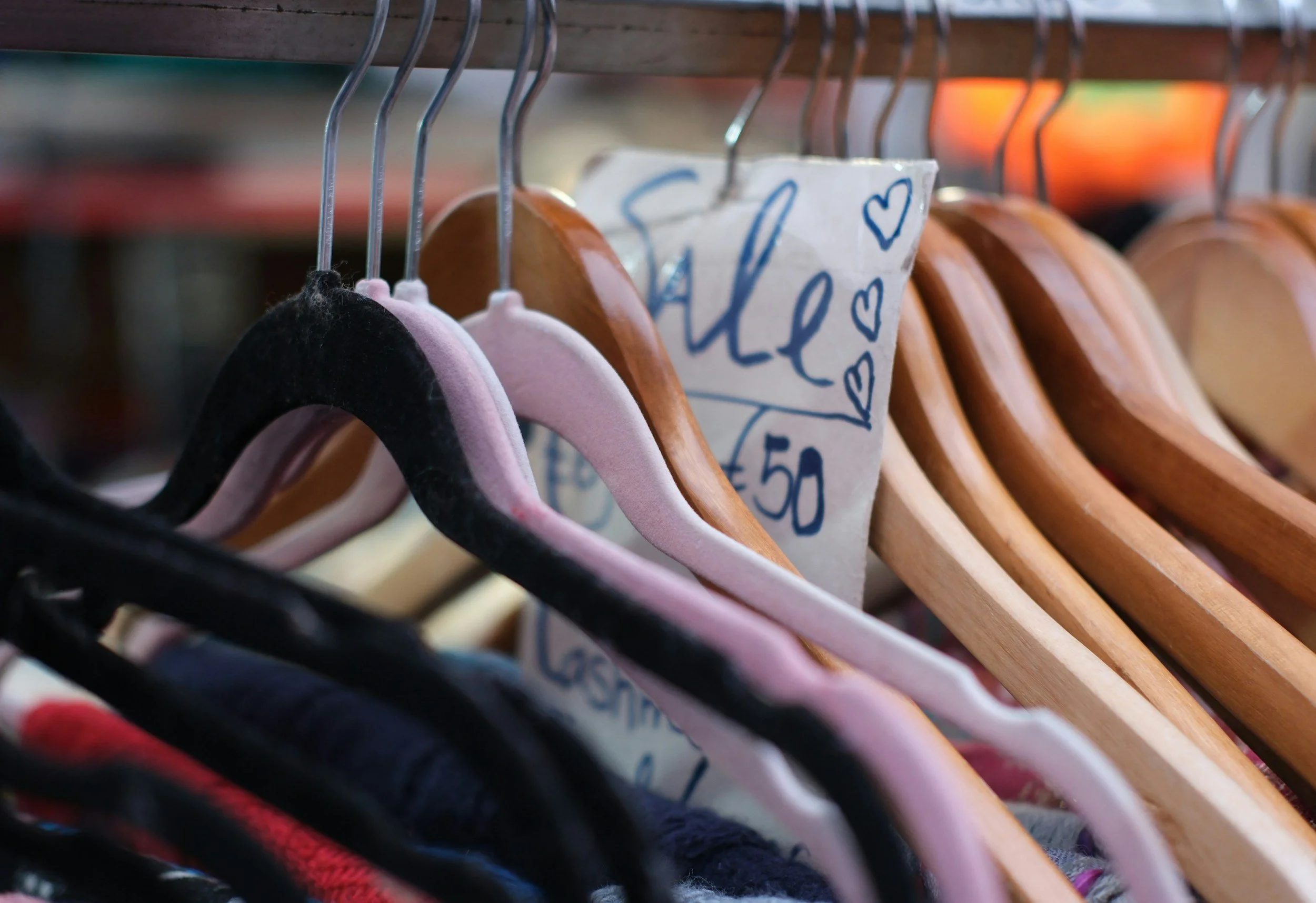 Resale clothes hang on hangers in a retail store, including velvety black and pink hangers and wooden hangers with a handwritten sign that reads 'Sale' decorated with hearts.