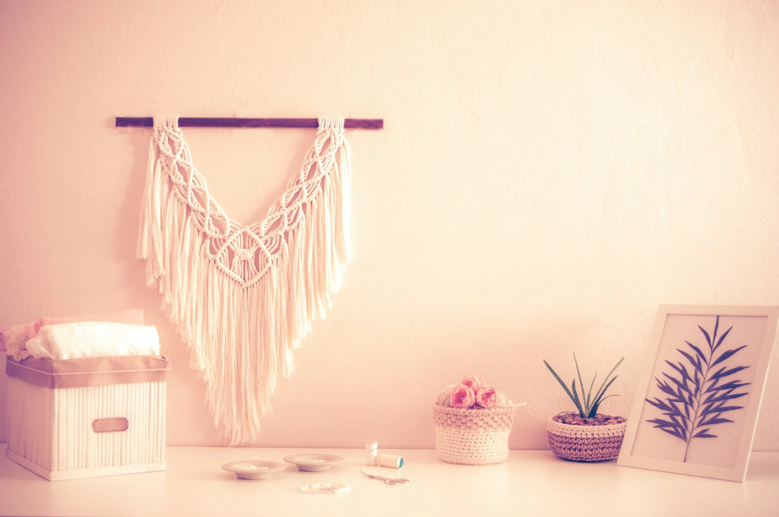 Decorative Macrame wall hanging with cream-colored fringe over a neutral wall. Small items including a box with tissue, pink roses in a woven basket, a potted plant, and framed botanical art on a white surface.