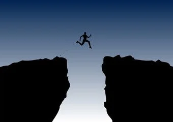 Silhouette of a person jumping from one cliff to another against a darkening sky.