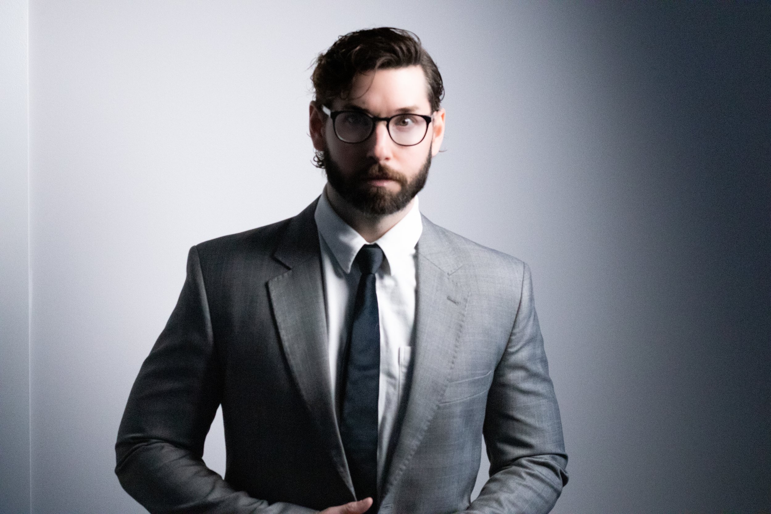 A man with dark hair and glasses wearing a gray suit, white shirt, and black tie standing against a light gray wall.