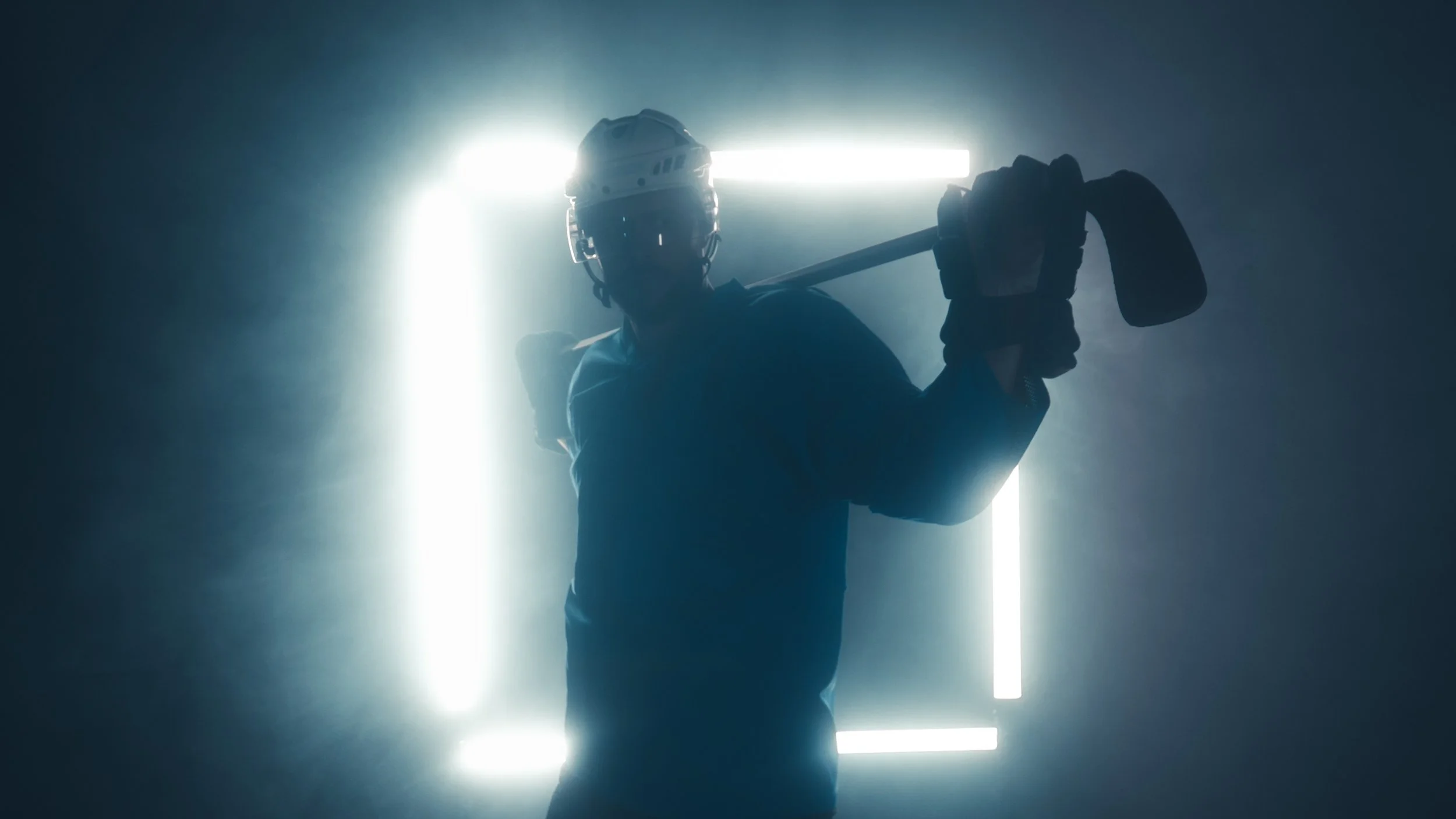 Silhouette of a person wearing a helmet and holding a sledgehammer against a bright light backlit background with neon square outline.