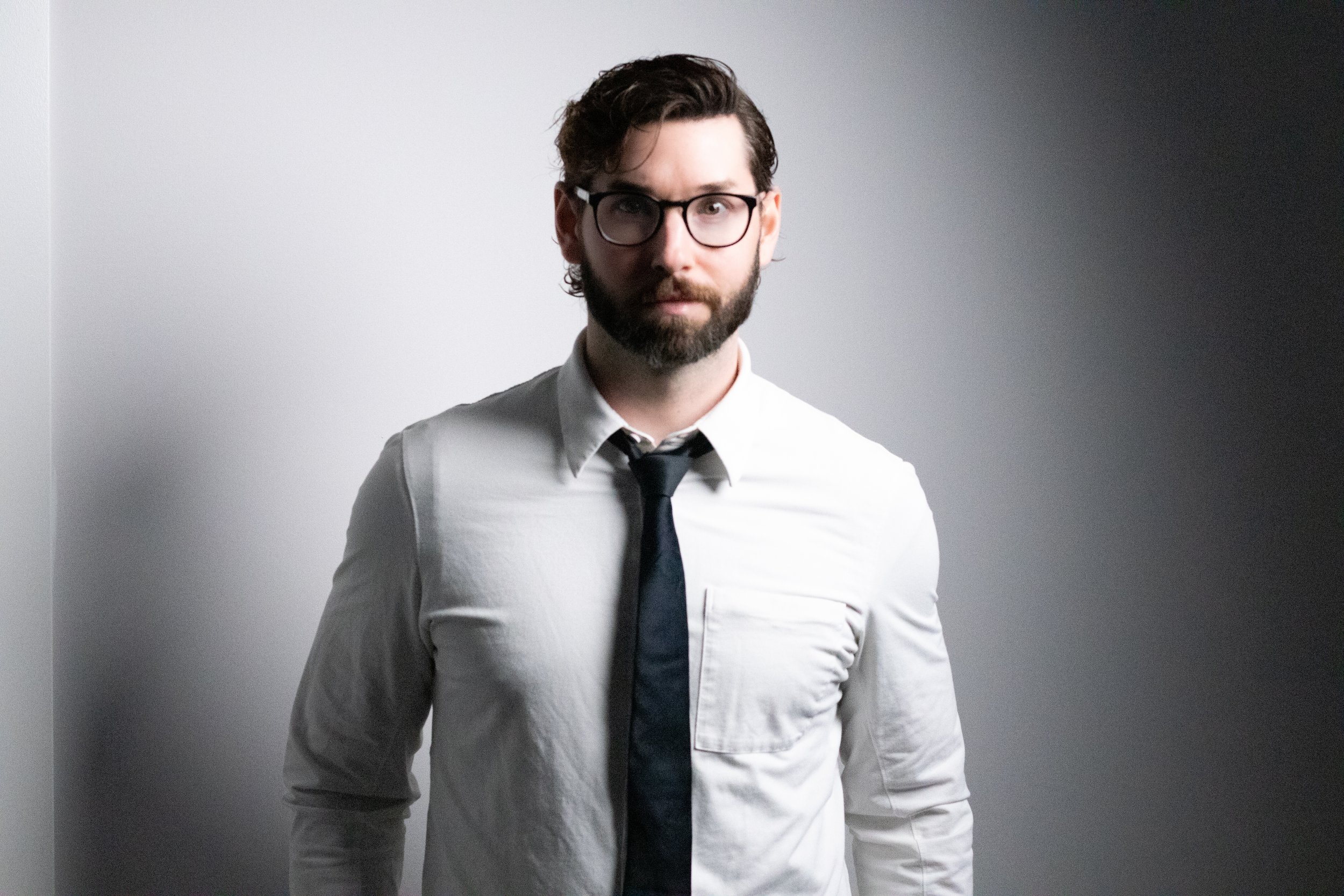 A man with glasses, dark hair, and a beard, wearing a white shirt and black tie, standing against a gray and white background.