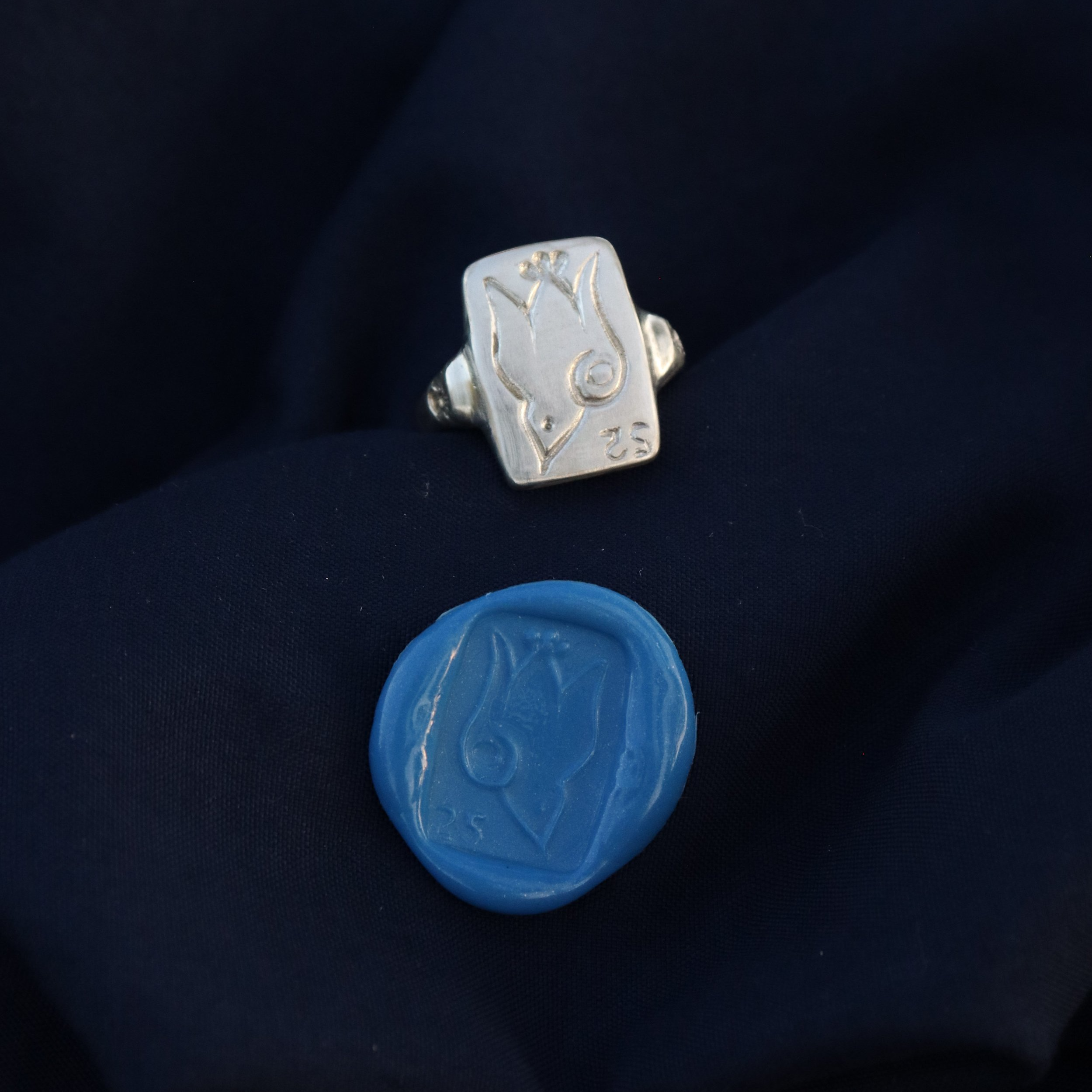 A silver ring with a bird engraving, and a blue wax seal with the same bird design, both resting on navy blue fabric.