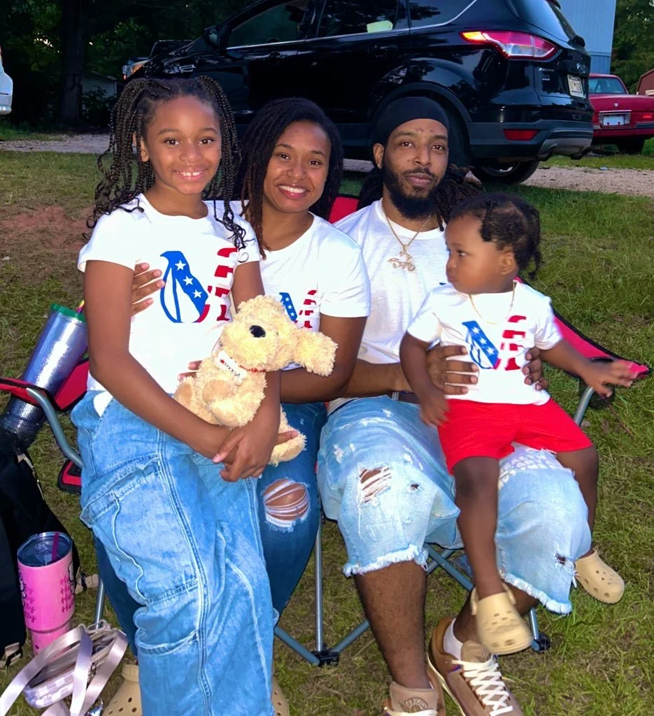 A family of four sitting on a camping chair outdoors, wearing matching white shirts with red, white, and blue designs. The girl on the left is holding a plush dog toy, the woman is smiling, the man has a beard and is wearing a black head cover, and t
