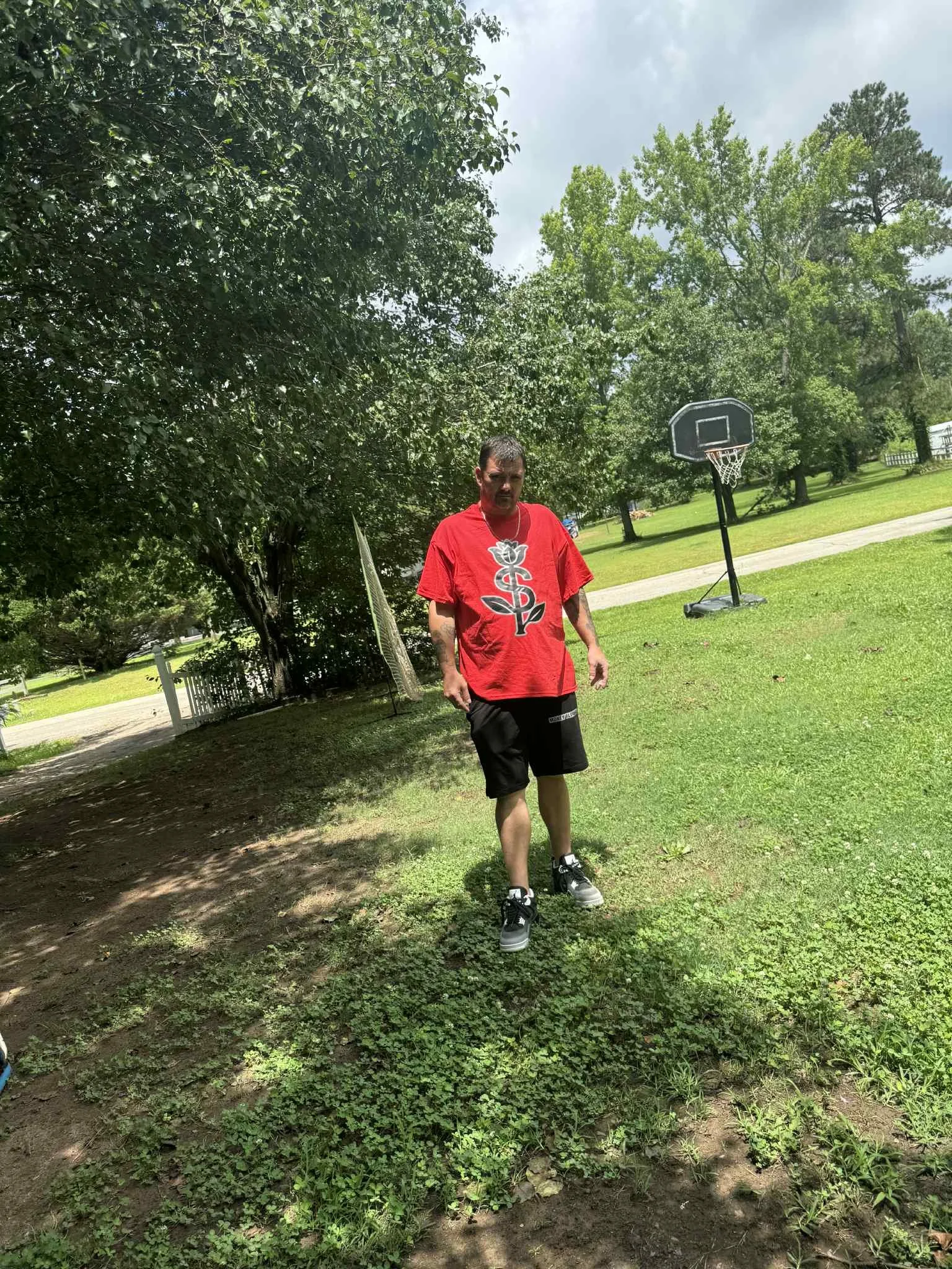 A man with a mustache and beard wearing a red shirt with black shorts and sneakers, standing outdoors on a grassy yard with trees and a basketball hoop in the background.