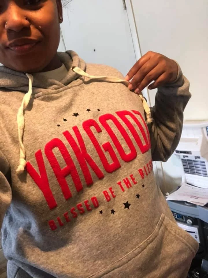 Person wearing a gray hoodie with red text reading 'YANKEE DIVIDED BE THE BEAT' and black stars, pointing to the hoodie. The person is smiling slightly, standing indoors near a printer.