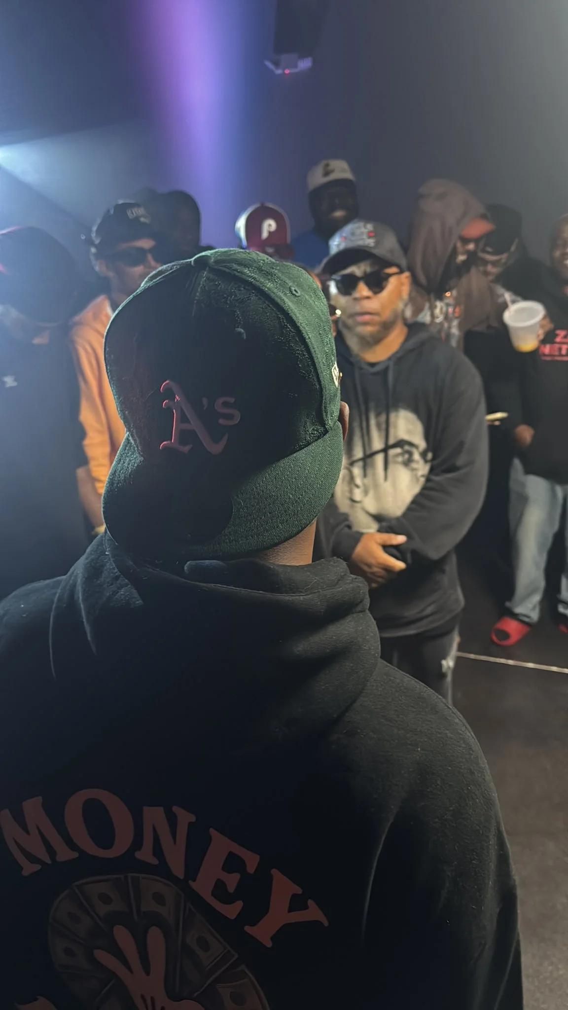 A group of people, mostly wearing hoodies and sunglasses, gathered indoors with a dark background and purple lighting, facing towards the camera.