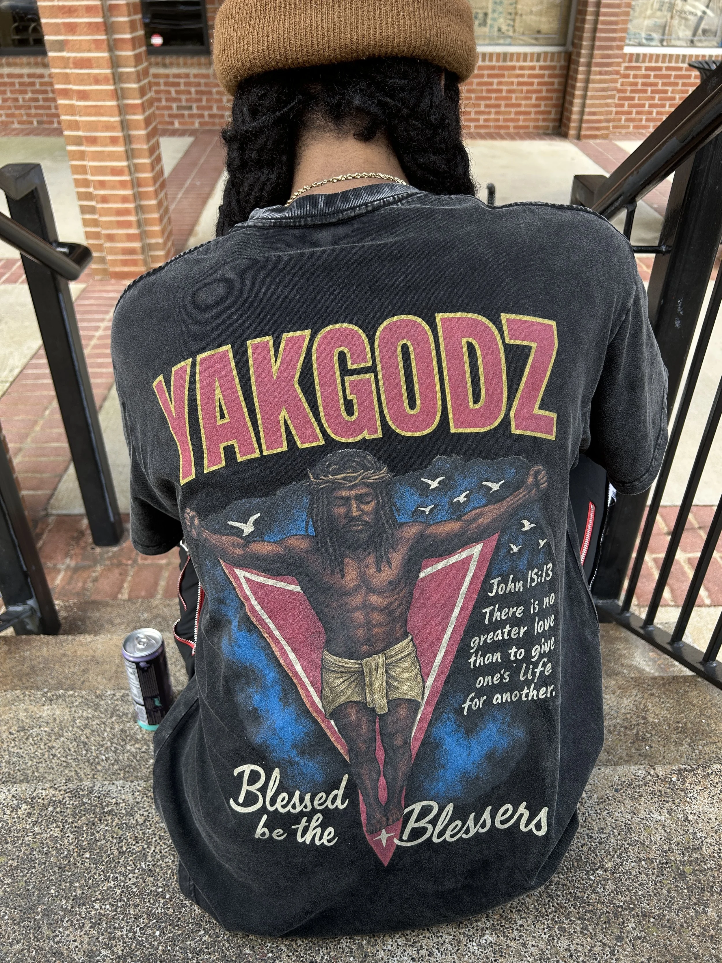Back of person wearing a black T-shirt with a graphic of Jesus on a cross and a Bible verse, standing outside near brick building and metal railing.