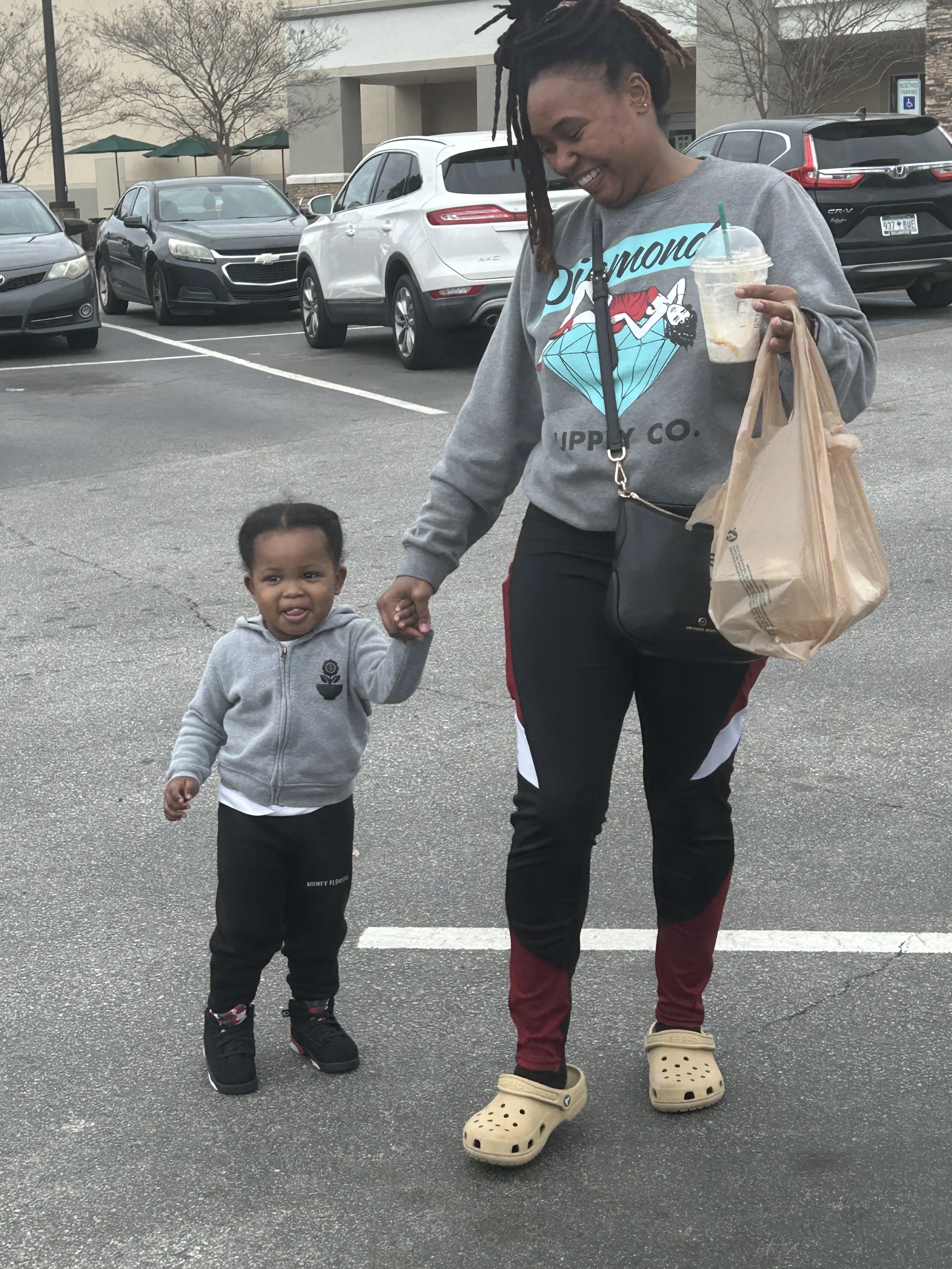 A woman and young child walking in a parking lot, holding hands. The woman is smiling, wearing a gray hoodie with a colorful graphic, black and red sweatpants, and beige Crocs. The child is wearing a gray jacket, black pants, and black sneakers with 