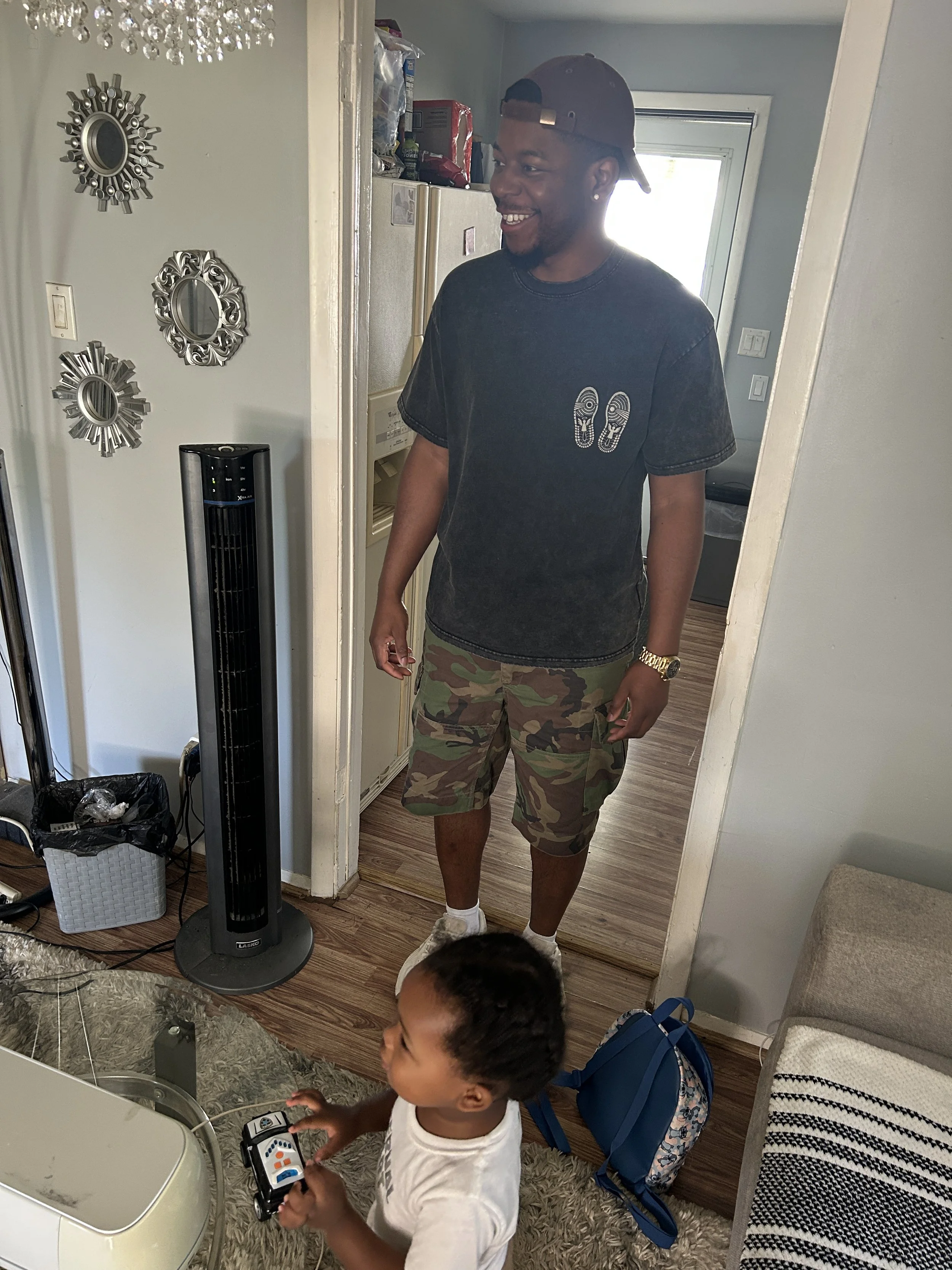 A young man wearing a brown cap, black t-shirt with shoe prints, and camouflage shorts stands in a kitchen near a mirror, smiling. A young boy with a device stands in front of a mirror, holding two remote controls. The room has decorative mirrors, a 