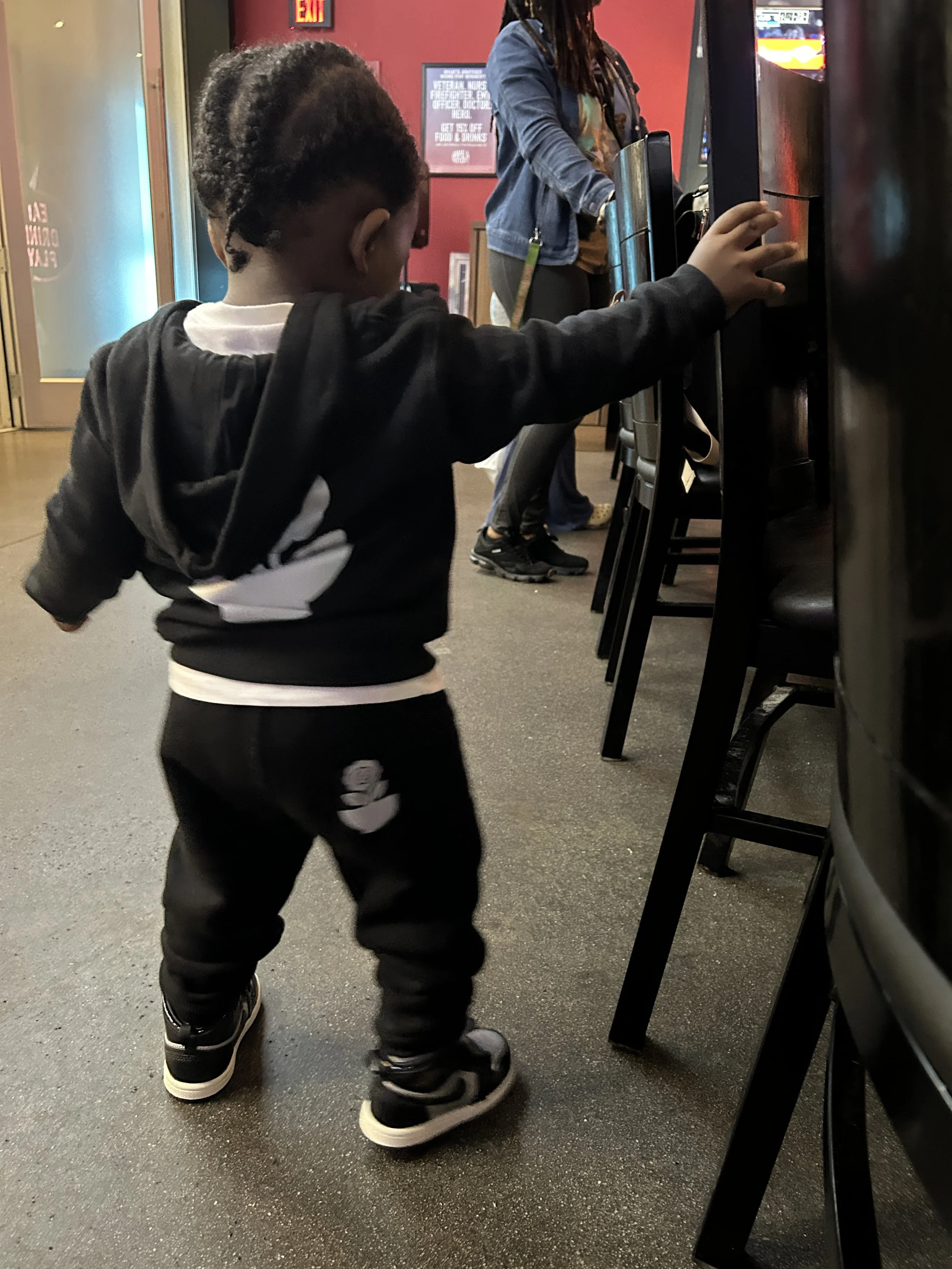 A young boy wearing black clothing and sneakers standing in front of an arcade machine, reaching out with his right hand.