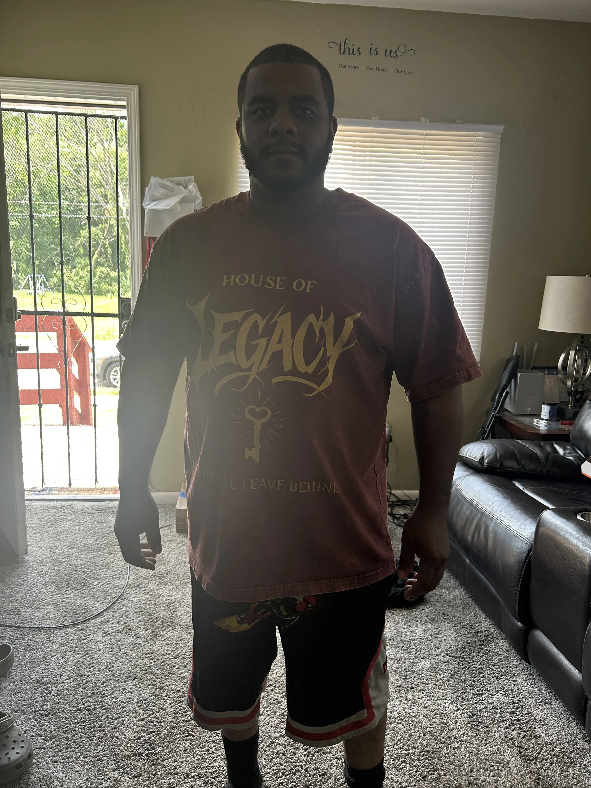A young man standing indoors in front of windows with blinds, wearing a maroon T-shirt with a graphic design, shorts, and socks. The room contains a black leather couch, a side table with items, and a lamp. The back door with a metal security screen 