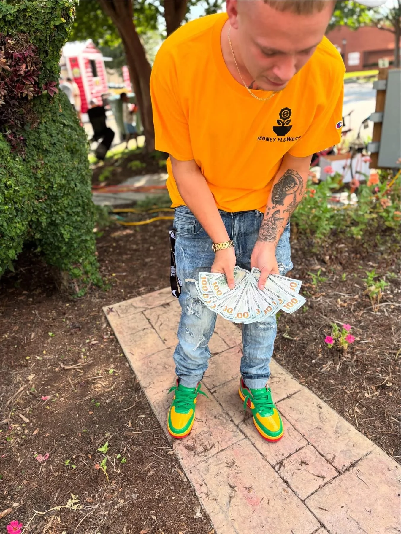 A young man wearing a yellow t-shirt, distressed jeans, and colorful sneakers stands outdoors on a brick pathway, holding multiple hundred-dollar bills fanned out in his hands.